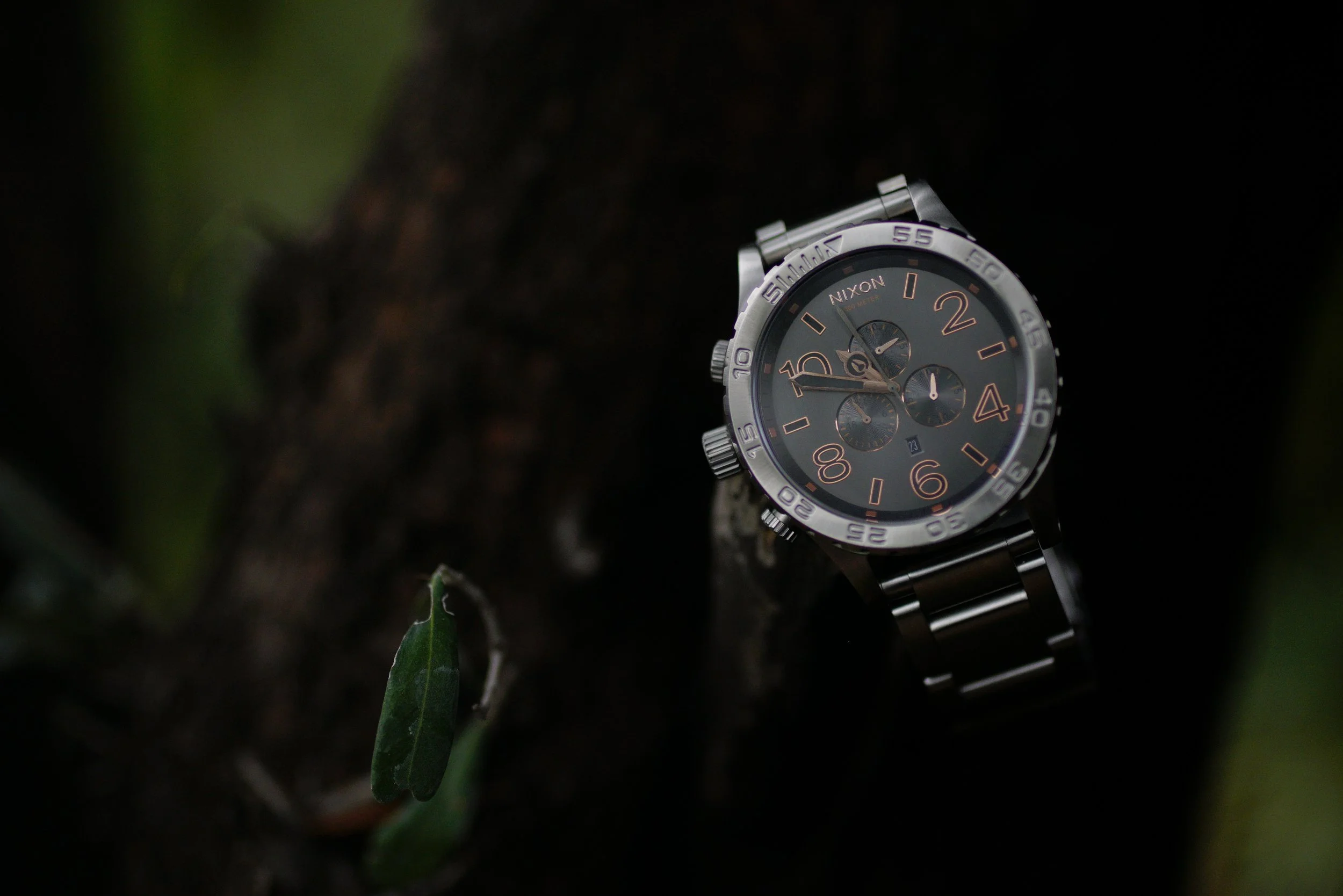 A silver wristwatch with a black face and rose gold accents, placed on a dark surface with blurred green leaves in the background.
