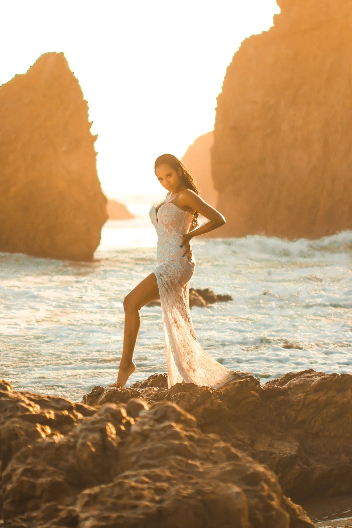 A woman in a lace dress standing on rocks at the beach during sunset, with large rock formations in the background.