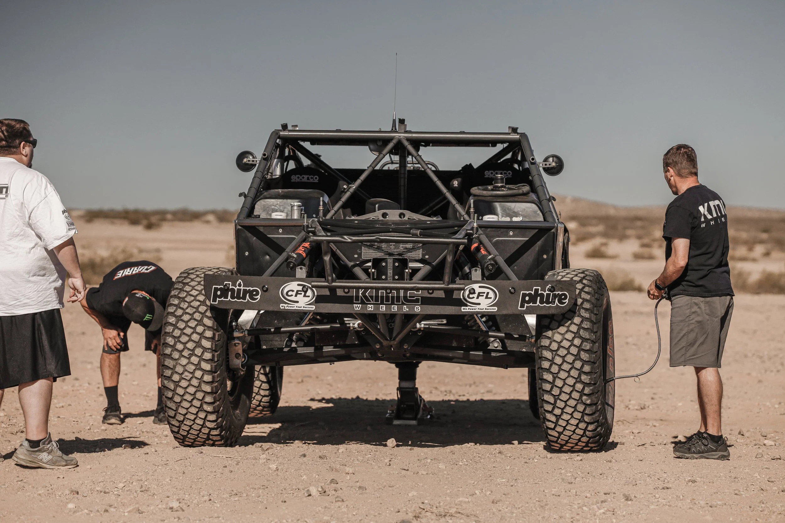 People working on a desert off-road racing vehicle with large tires and a visible chassis, in a sandy, open landscape under clear skies.