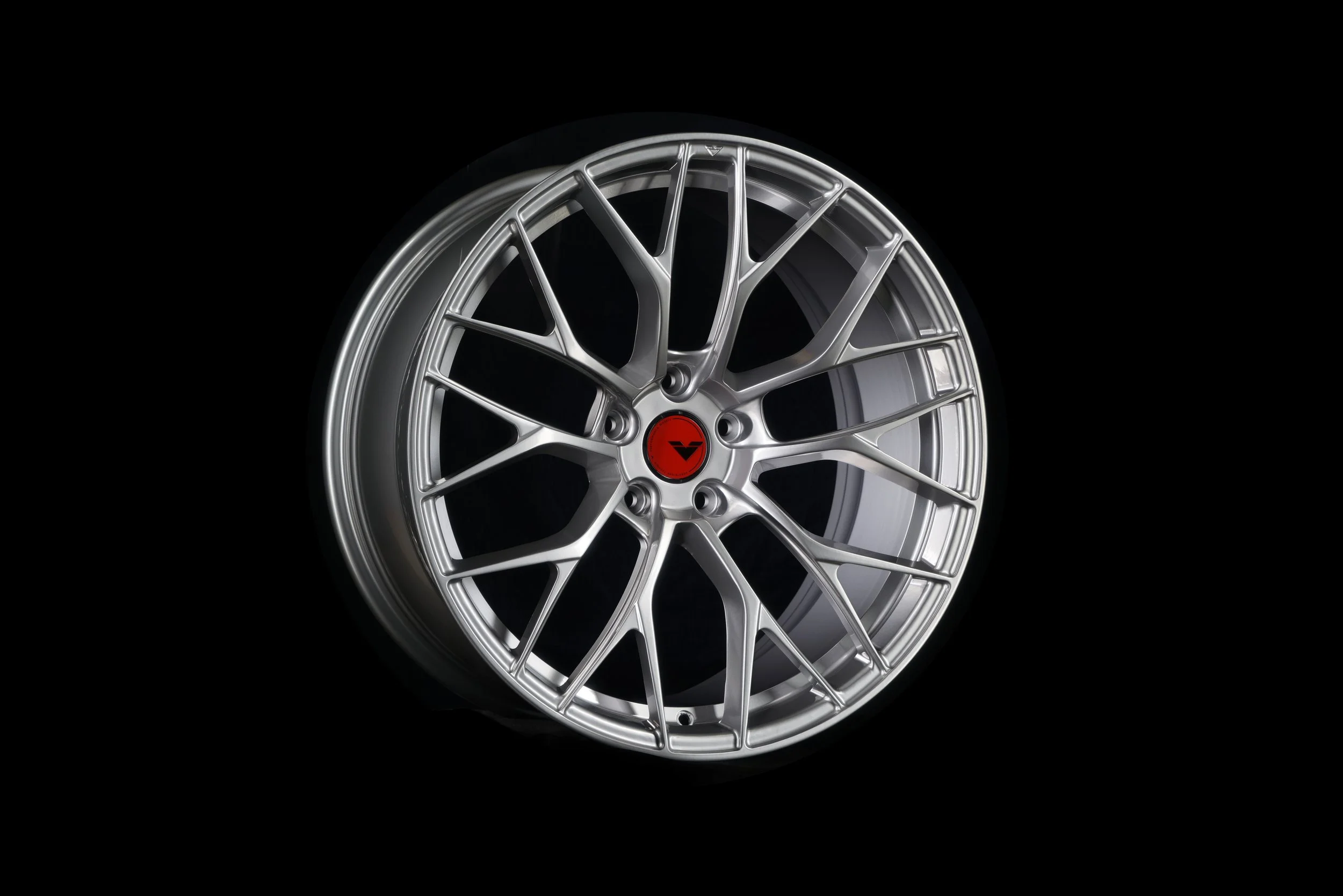 A close-up of a silver alloy wheel with an intricate spoke design, centered on a red hubcap with a black logo, set against a black background.