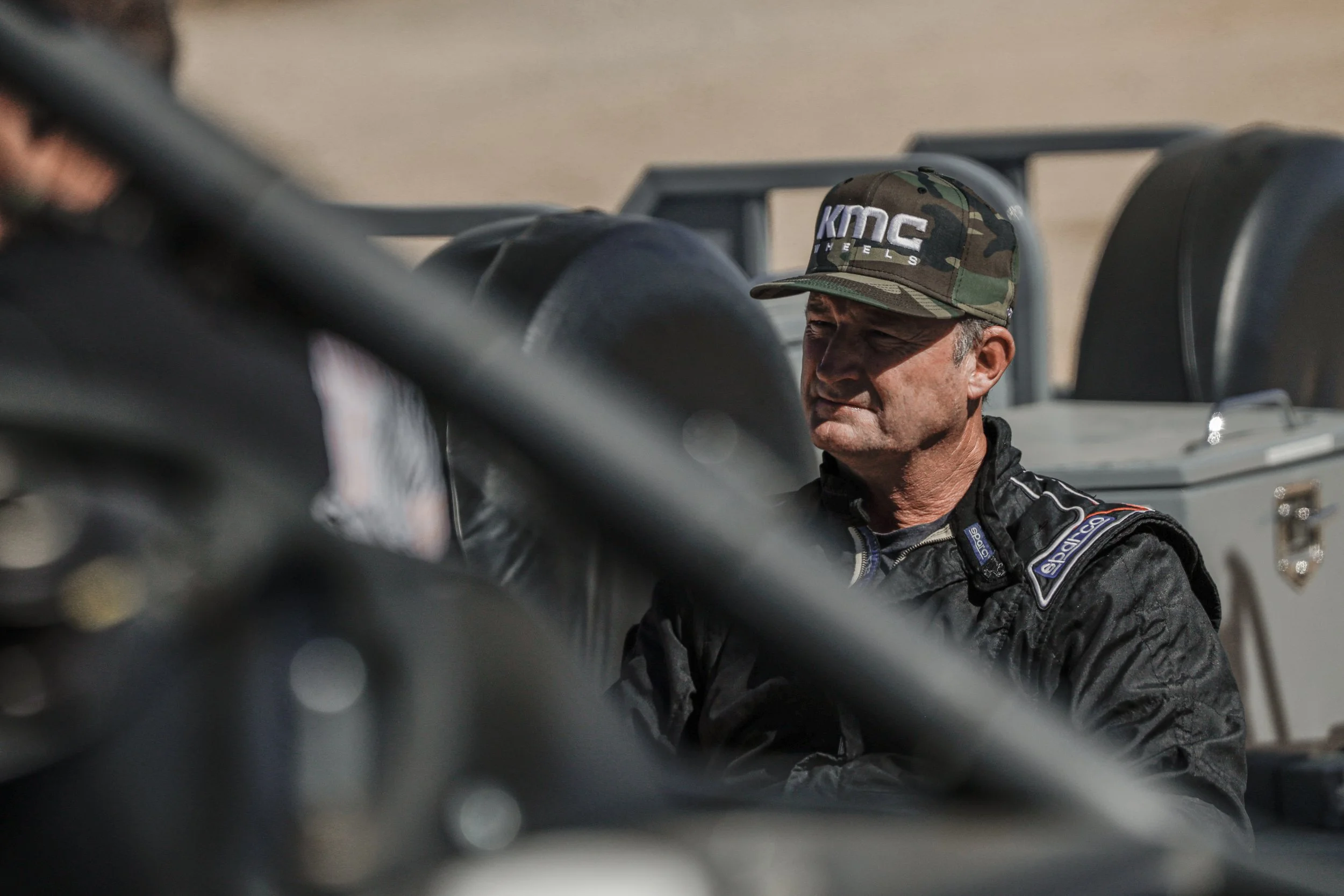 A man in a racing suit and a camouflage cap with 'KMC' logo, standing beside a racing car, in a desert or dry landscape.