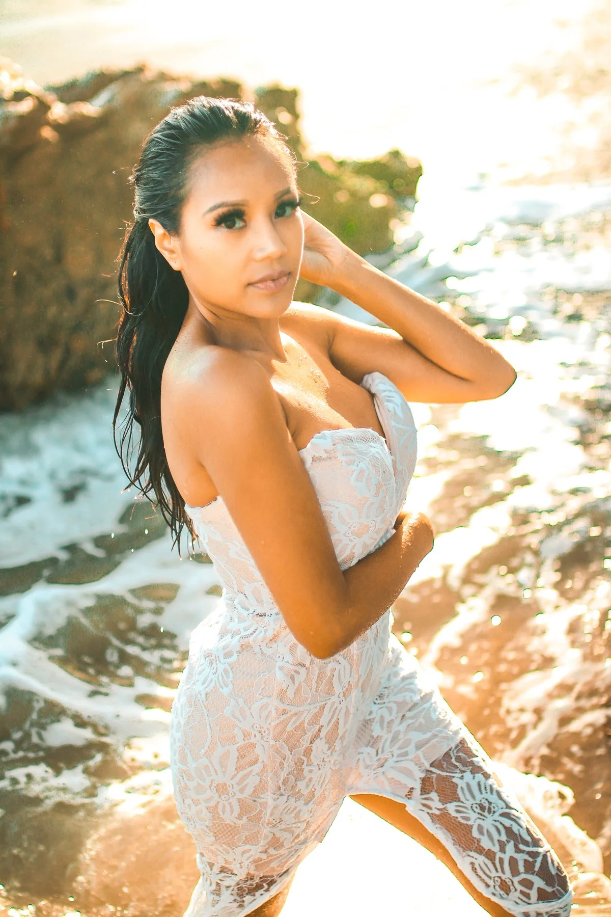 A woman in a white lace dress standing in shallow water on a beach at sunset.
