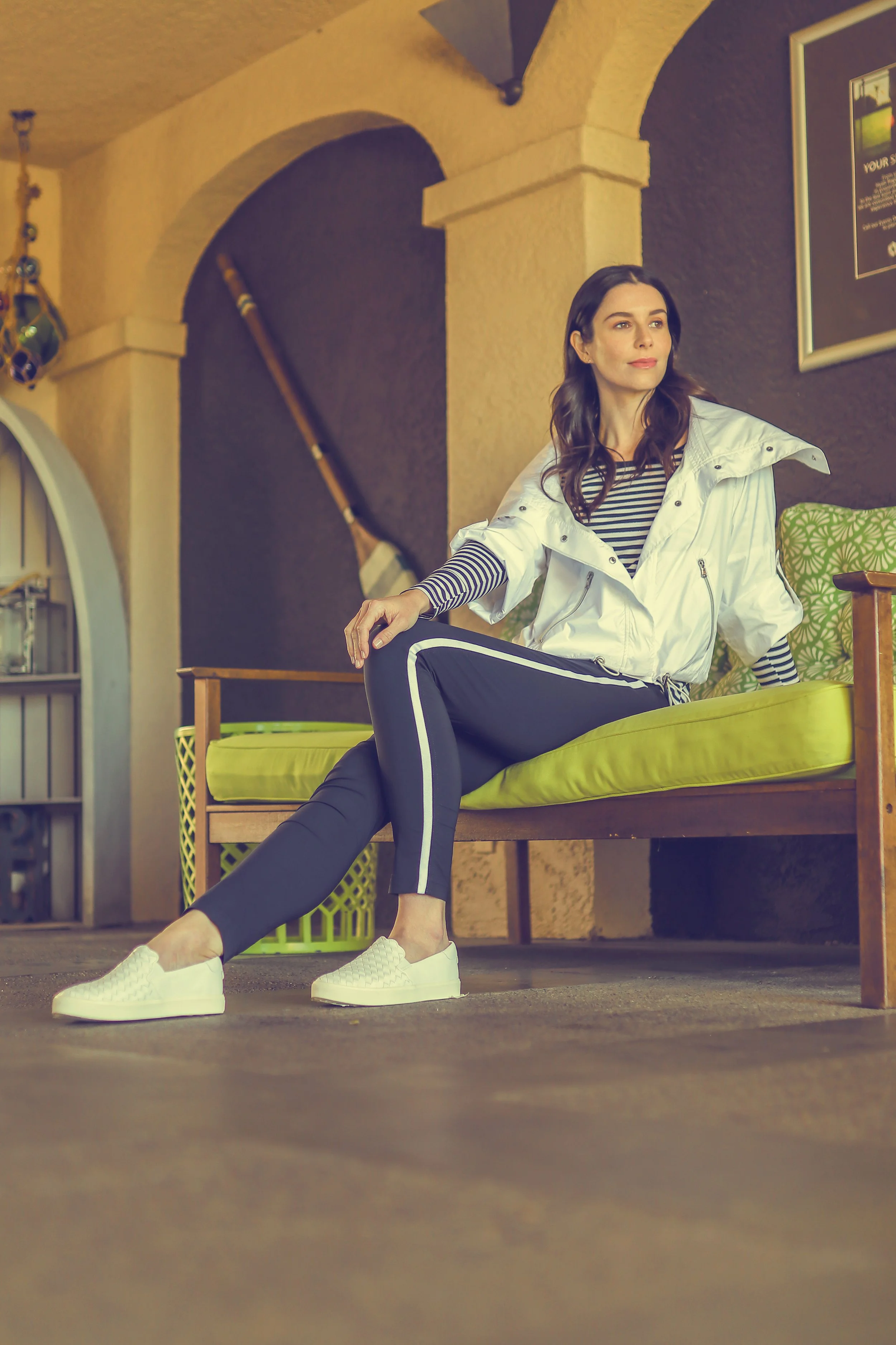 A woman sitting on a yellow cushioned outdoor sofa, wearing a white jacket, black athletic pants with white stripes, a striped shirt, and white sneakers, in a room with warm yellow walls, arched architectural details, and decorative items.