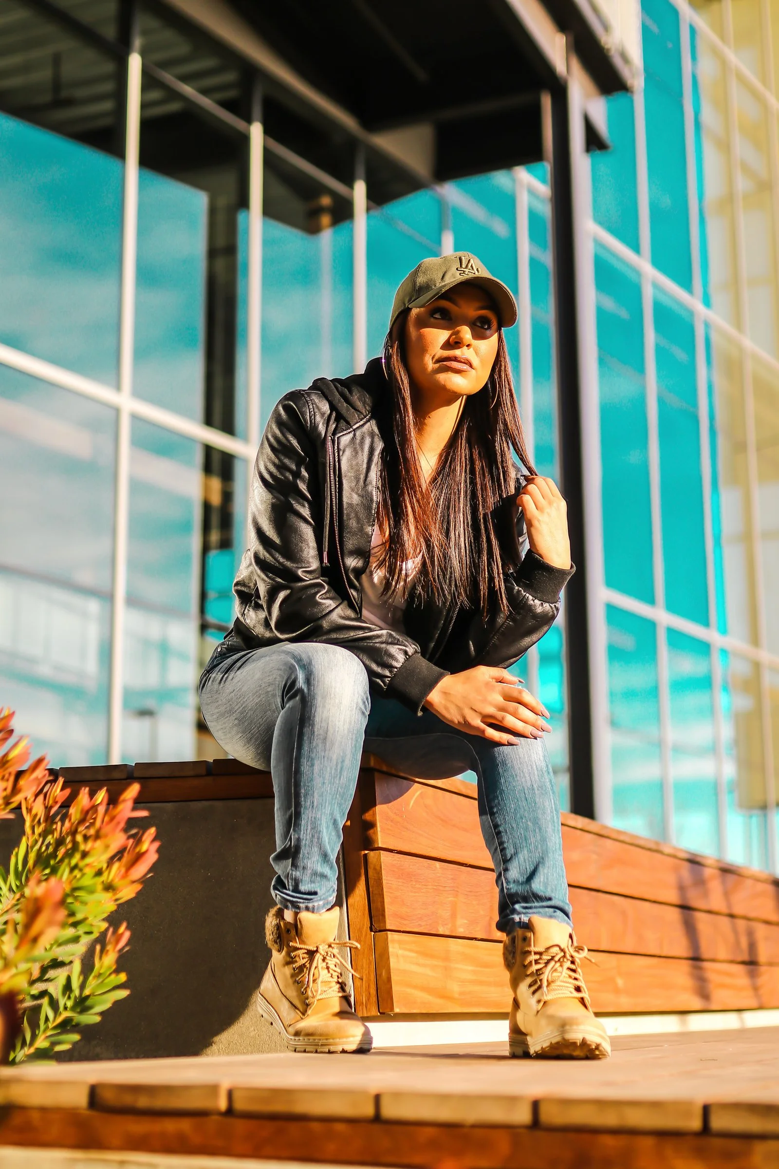 A young woman with long dark hair sitting on a wooden ledge outside a modern glass building, wearing a black leather jacket, gray jeans, tan boots, and a green baseball cap.