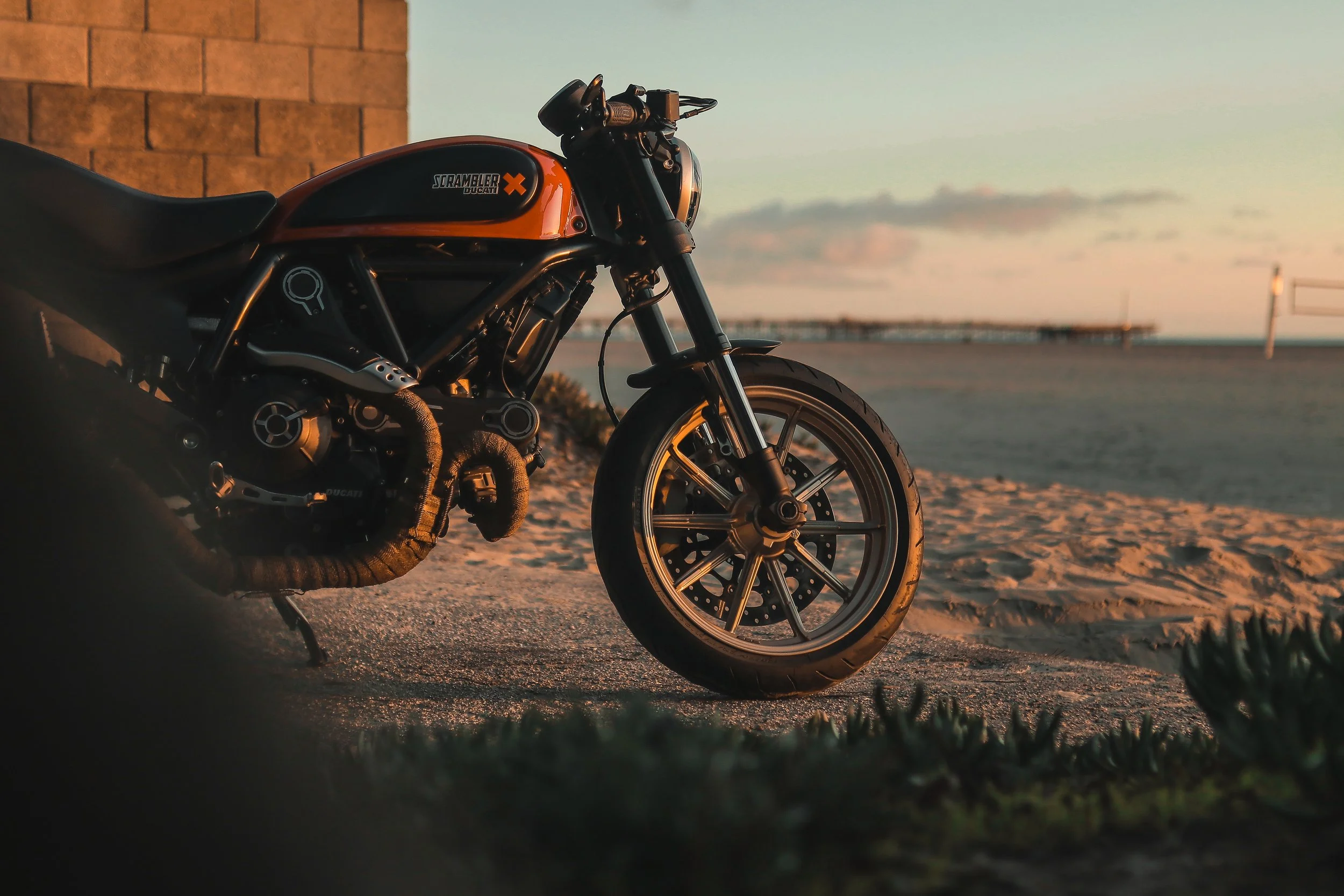A black and orange Ducati Scrambler motorcycle parked on a sandy beach during sunset, with a pier in the background.