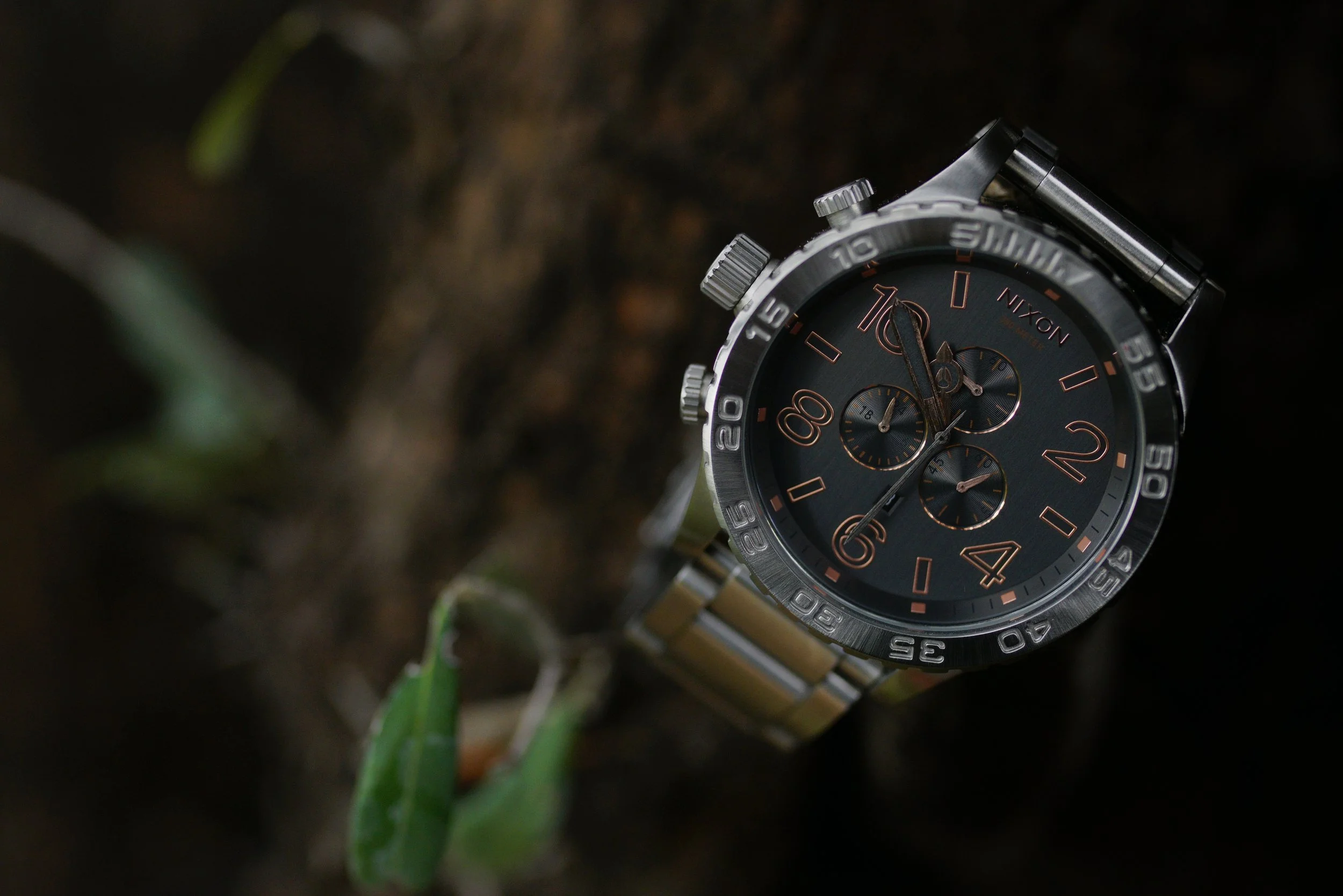 A close-up of a black Nixon wristwatch with orange accents, placed outdoors on soil with blurred green leaves in the foreground.