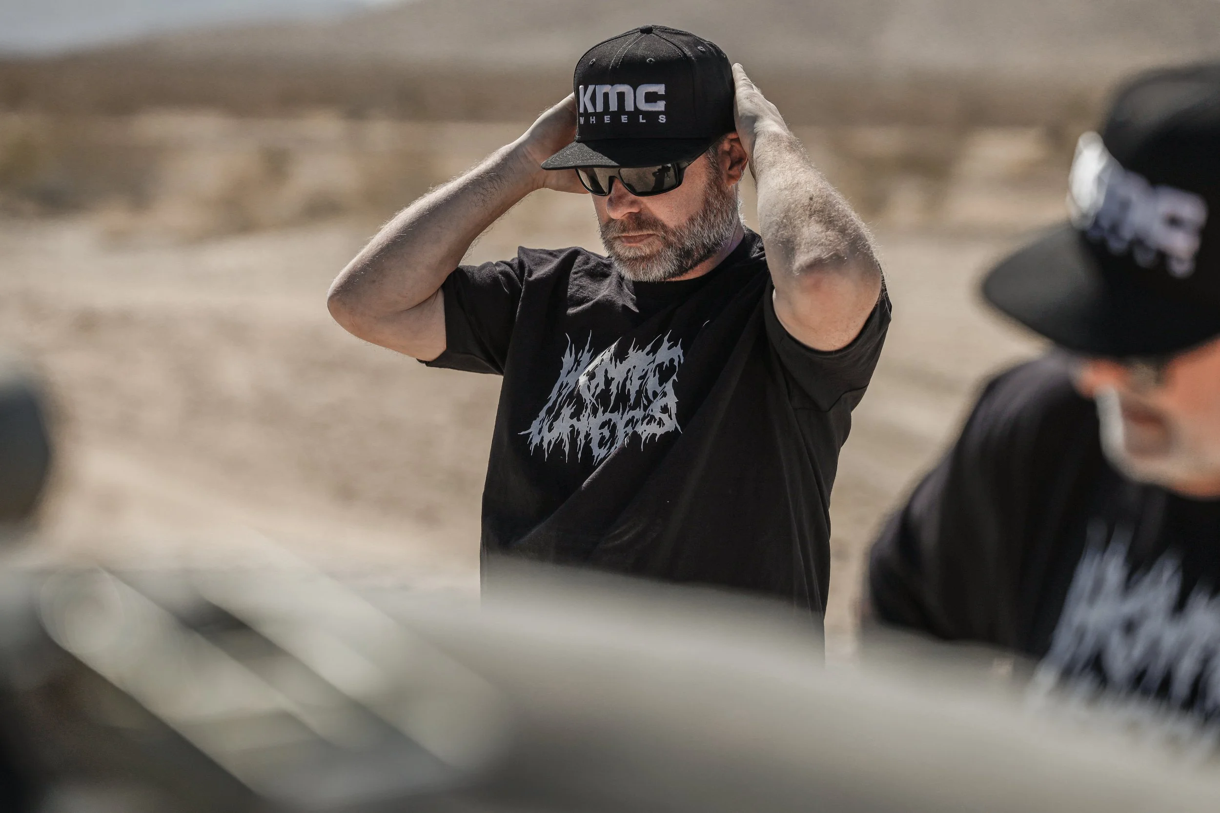 A man wearing sunglasses, a black cap with 'KMC' logo, and a black T-shirt with a white graphic design, adjusting his cap outdoors in a desert-like area.