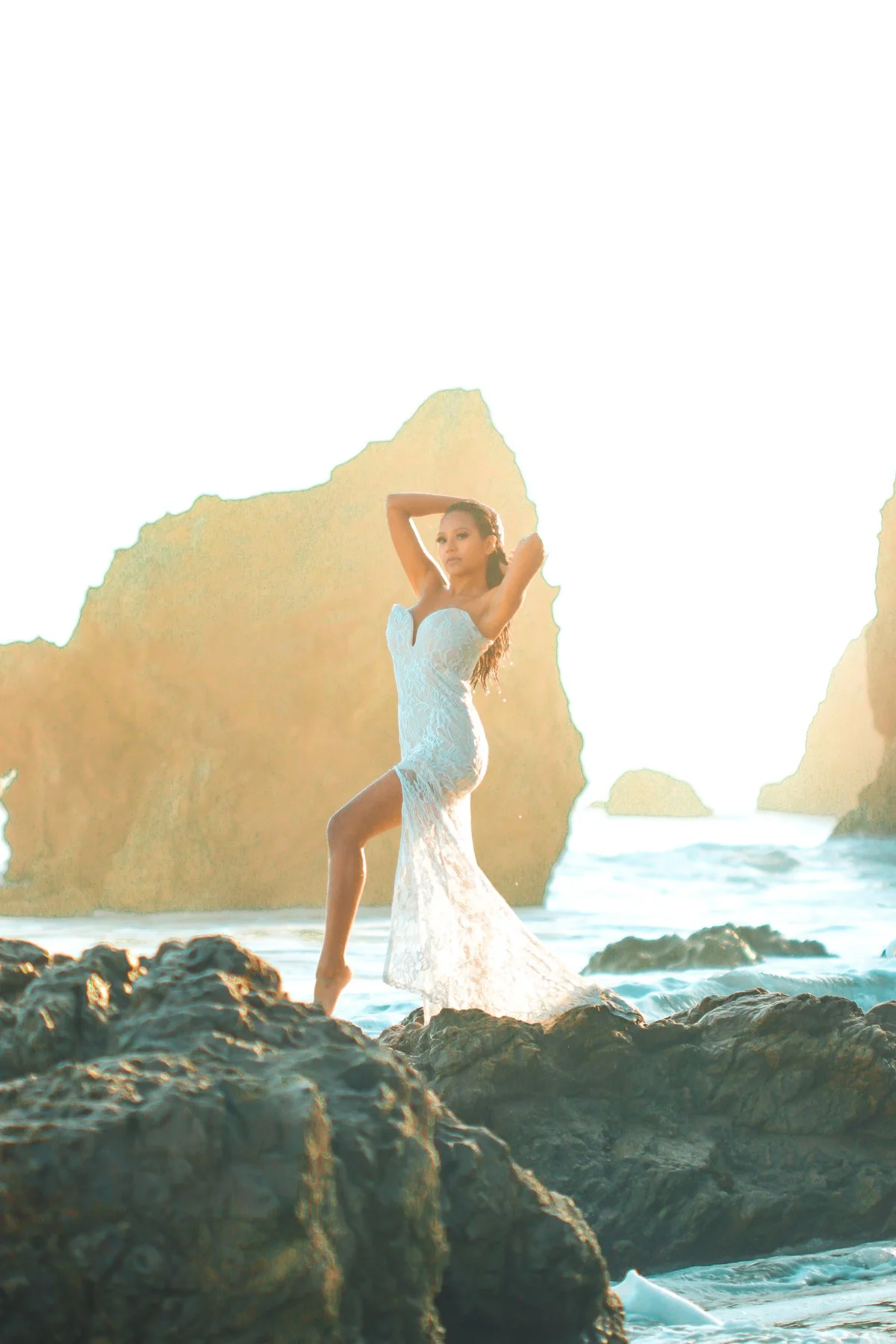 Woman in a white lace dress standing on rocks at the beach during sunrise or sunset, with large rock formations in the background.