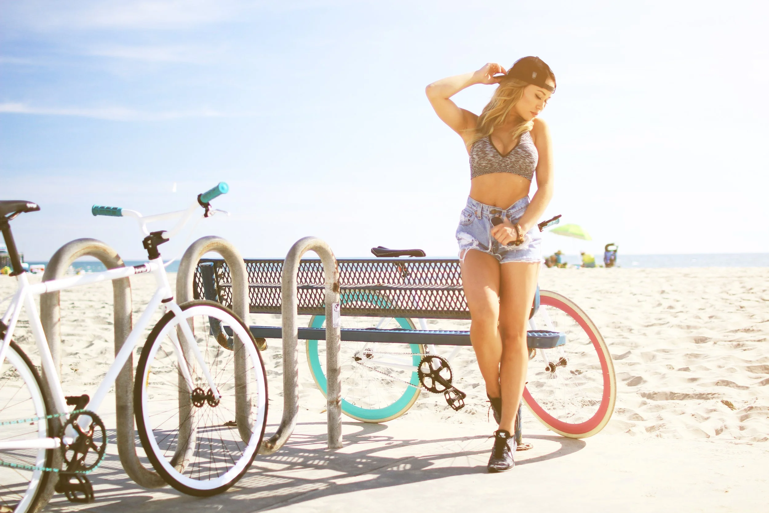 A woman with long blonde hair, wearing a black cap, a sports bra, and denim shorts, stands by bicycles on a beach with sand and ocean in the background.