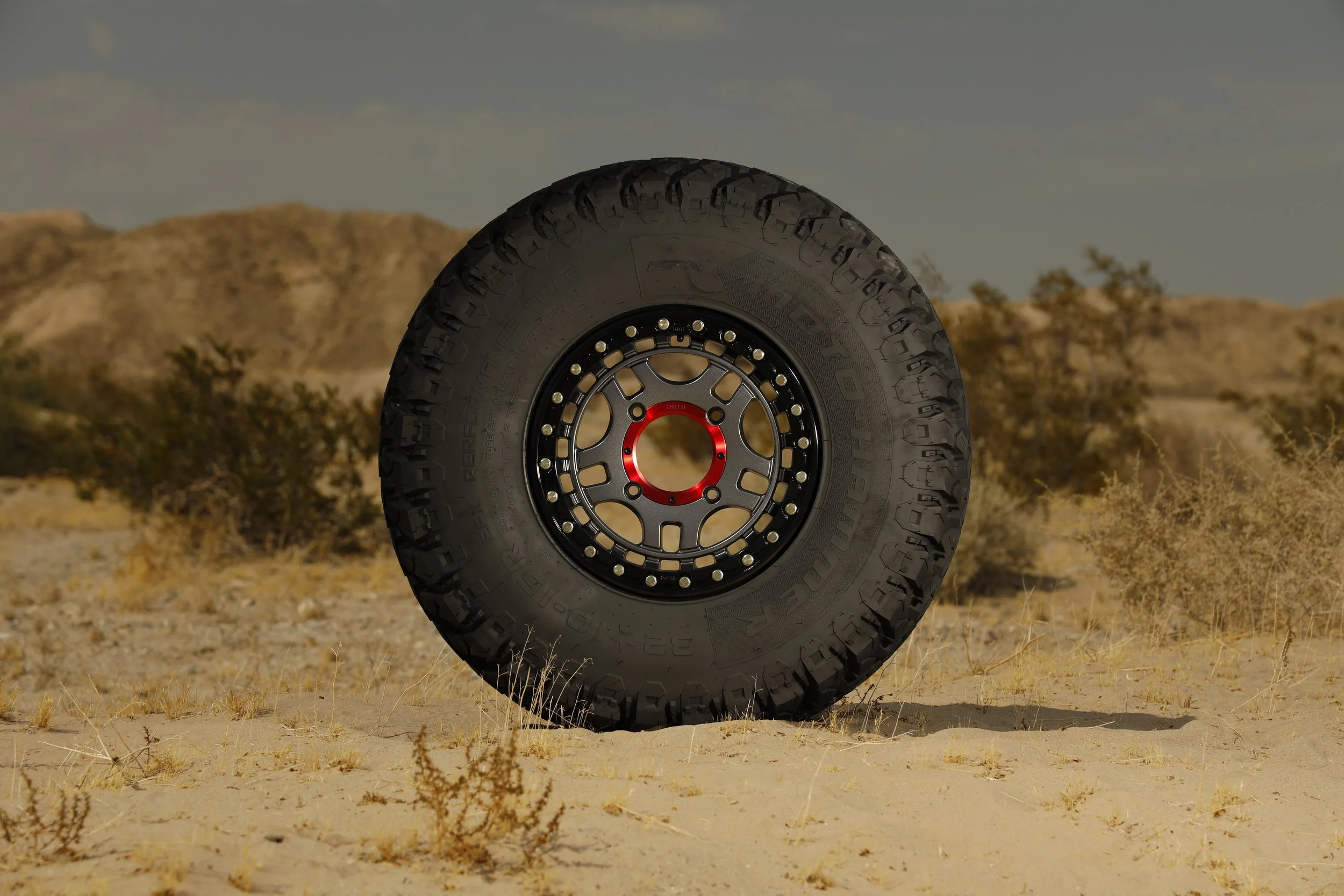 A close-up of a large off-road tire standing upright on sandy desert terrain with sparse shrubbery and distant hills under a partly cloudy sky.