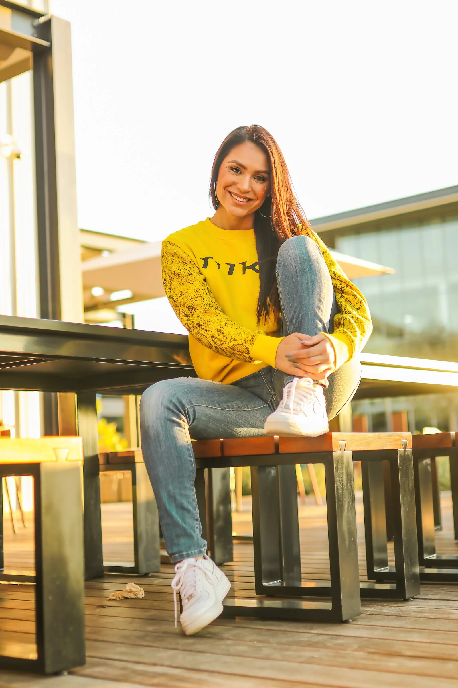 A young woman in a yellow sweatshirt and jeans sitting on a bench outdoors, smiling at the camera during sunset.