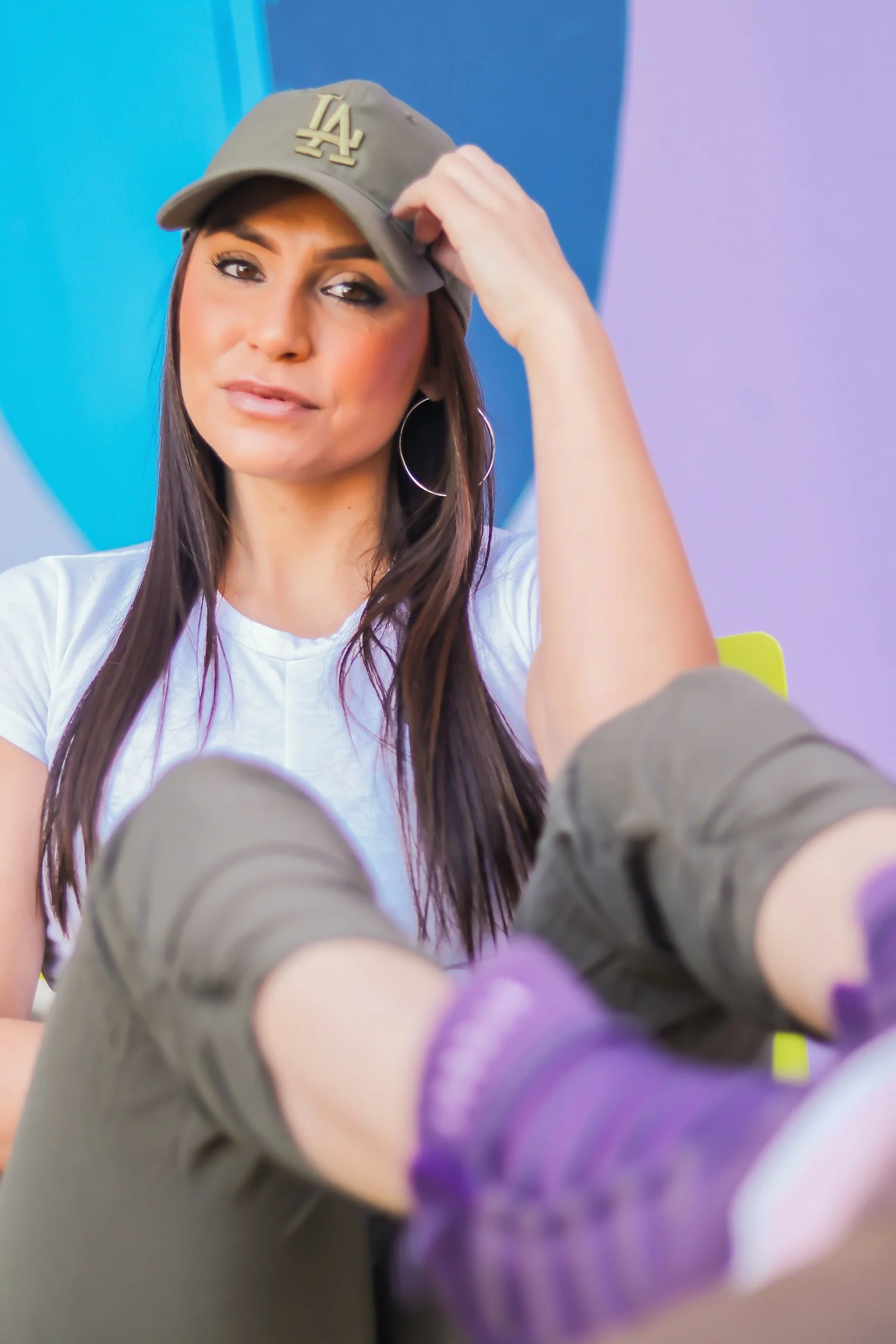 A woman wearing a beige LA baseball cap, white t-shirt, hoop earrings, and makeup, sitting in front of a colorful background, looking at the camera with a relaxed expression.