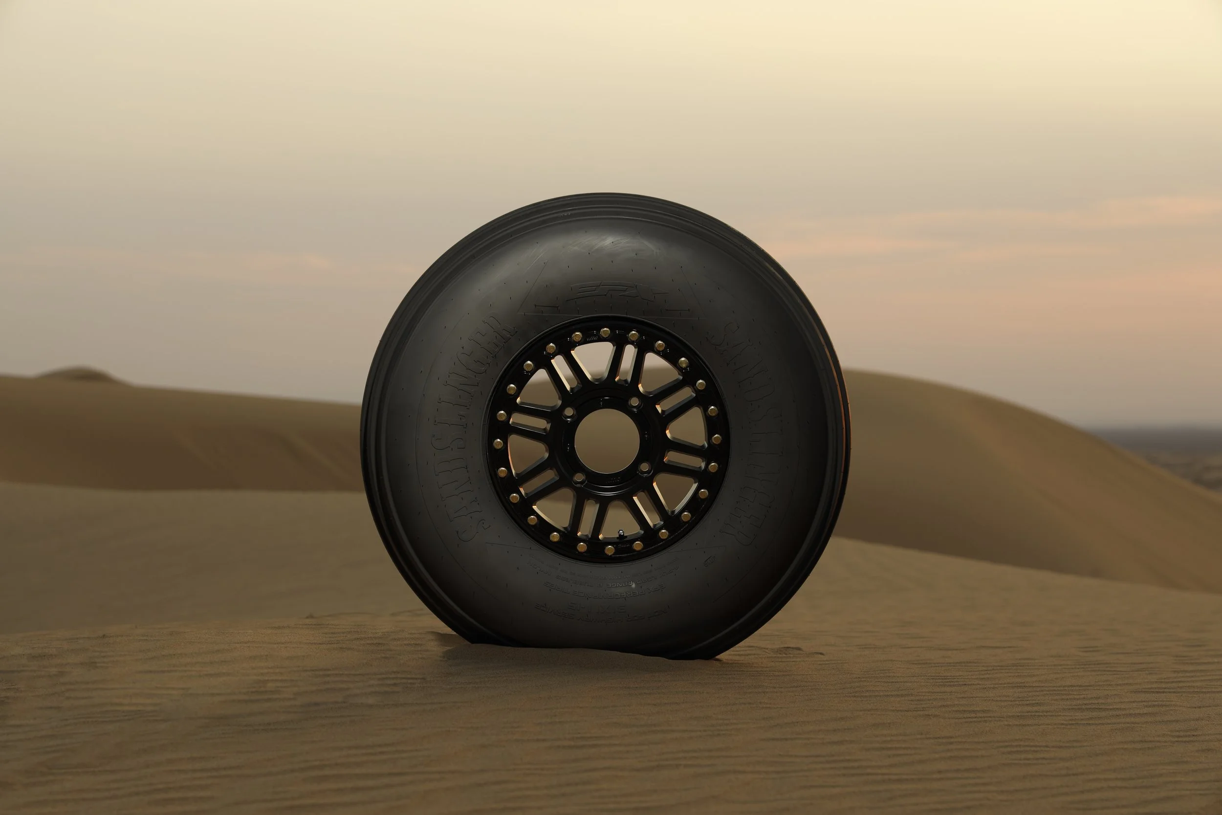 A vehicle tire in a desert landscape with sand dunes under a pastel-colored sky at sunset.