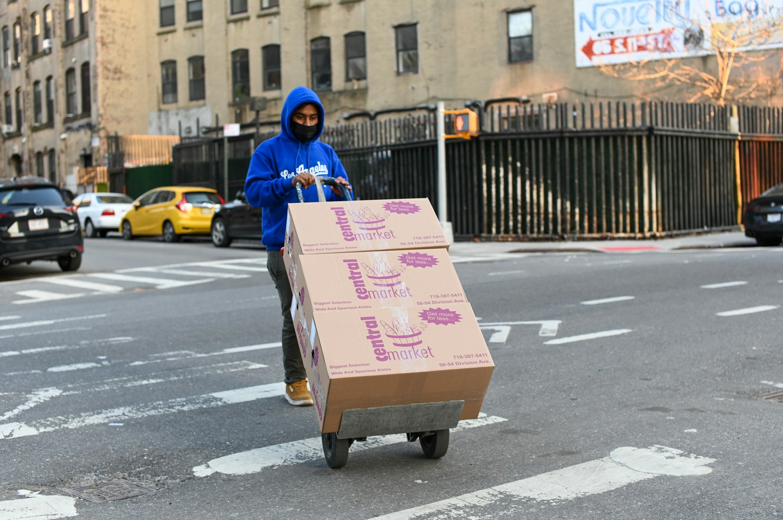 A person wearing a blue hoodie and black face mask pushes a hand truck loaded with three boxes labeled 'central market' across a city street at a crosswalk, with parked cars and an old building in the background.