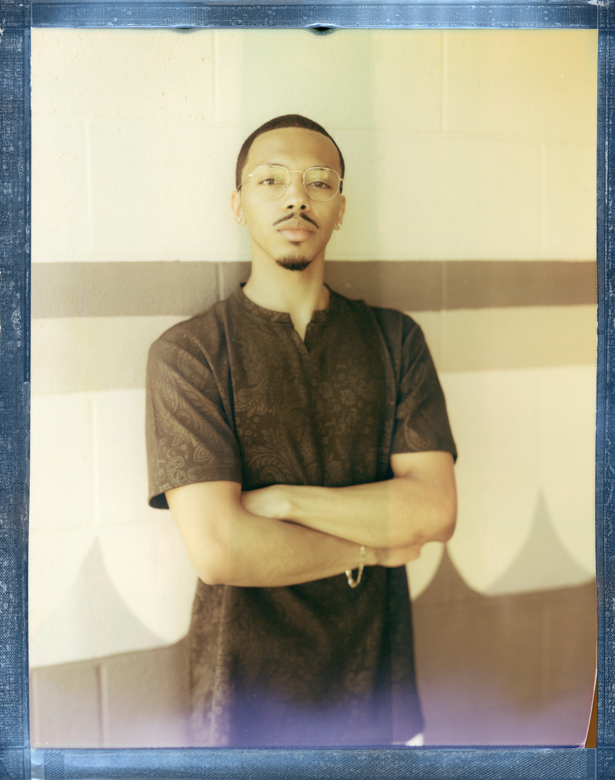Portrait of a young man with glasses and a goatee, standing against a striped wall with arms crossed, looking at the camera.