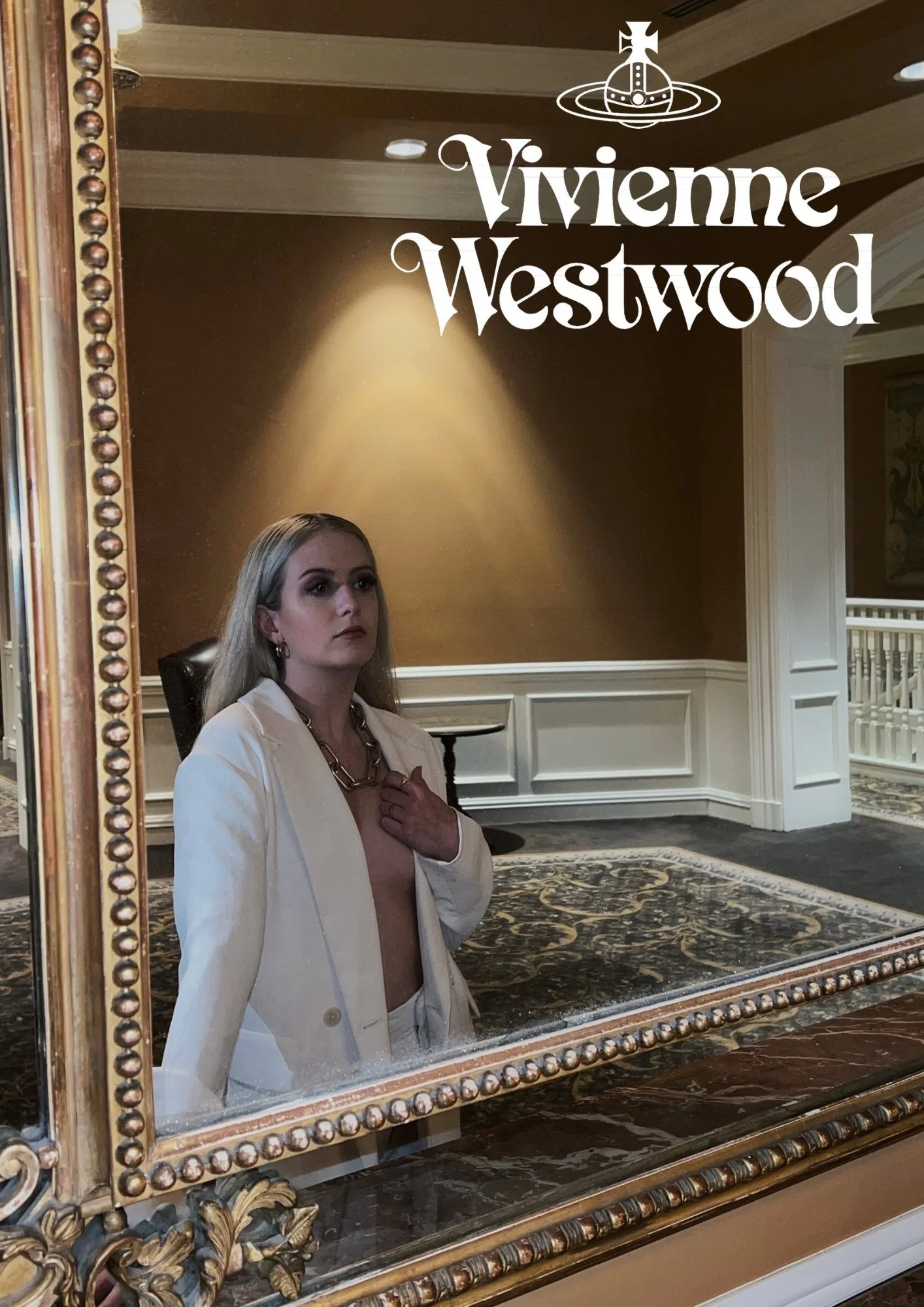 A woman in a beige blazer and matching pants is standing in front of a large mirror inside a luxurious room, with her hand on her chest. The room has ornate decor, including a patterned rug, wainscoting, and a dark brown wall. The mirror's frame feat