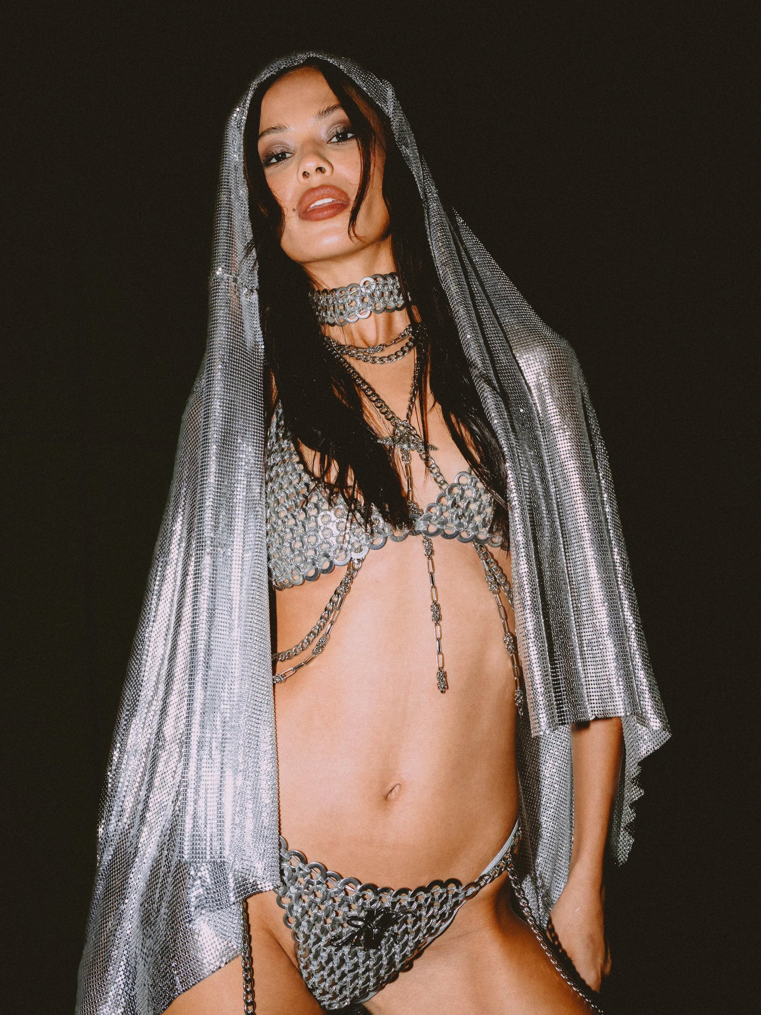 A woman with black hair and makeup, wearing multiple silver chain necklaces, a metallic silver sheer shawl, and silver chain lingerie, standing against a black background.