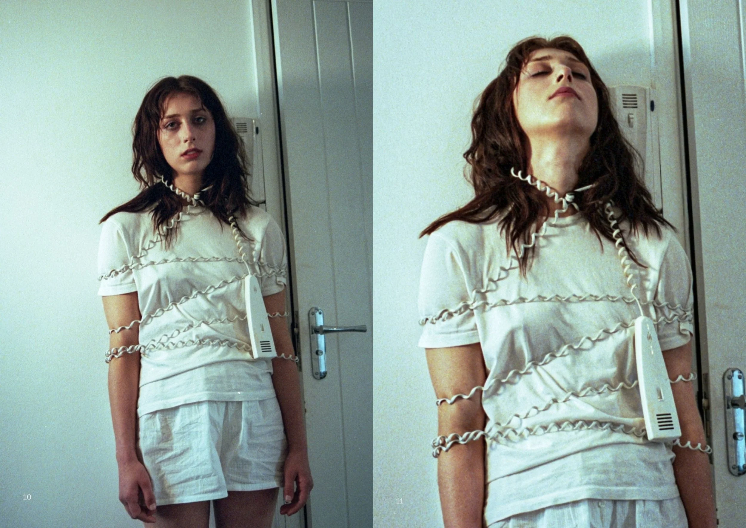 A woman with long, wavy dark hair standing in front of a mirror, looking at the camera with a neutral expression, wearing a white dress with black thread detailing and a choker, in a room with plain white walls and a door in the background.