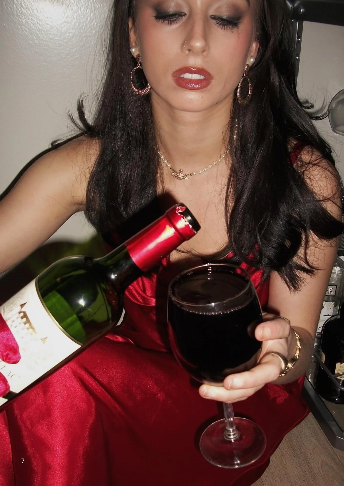 A woman with dark hair, makeup, and jewelry holding a glass of red wine. She is wearing a red dress and sitting at a table with a bottle of wine in front of her.