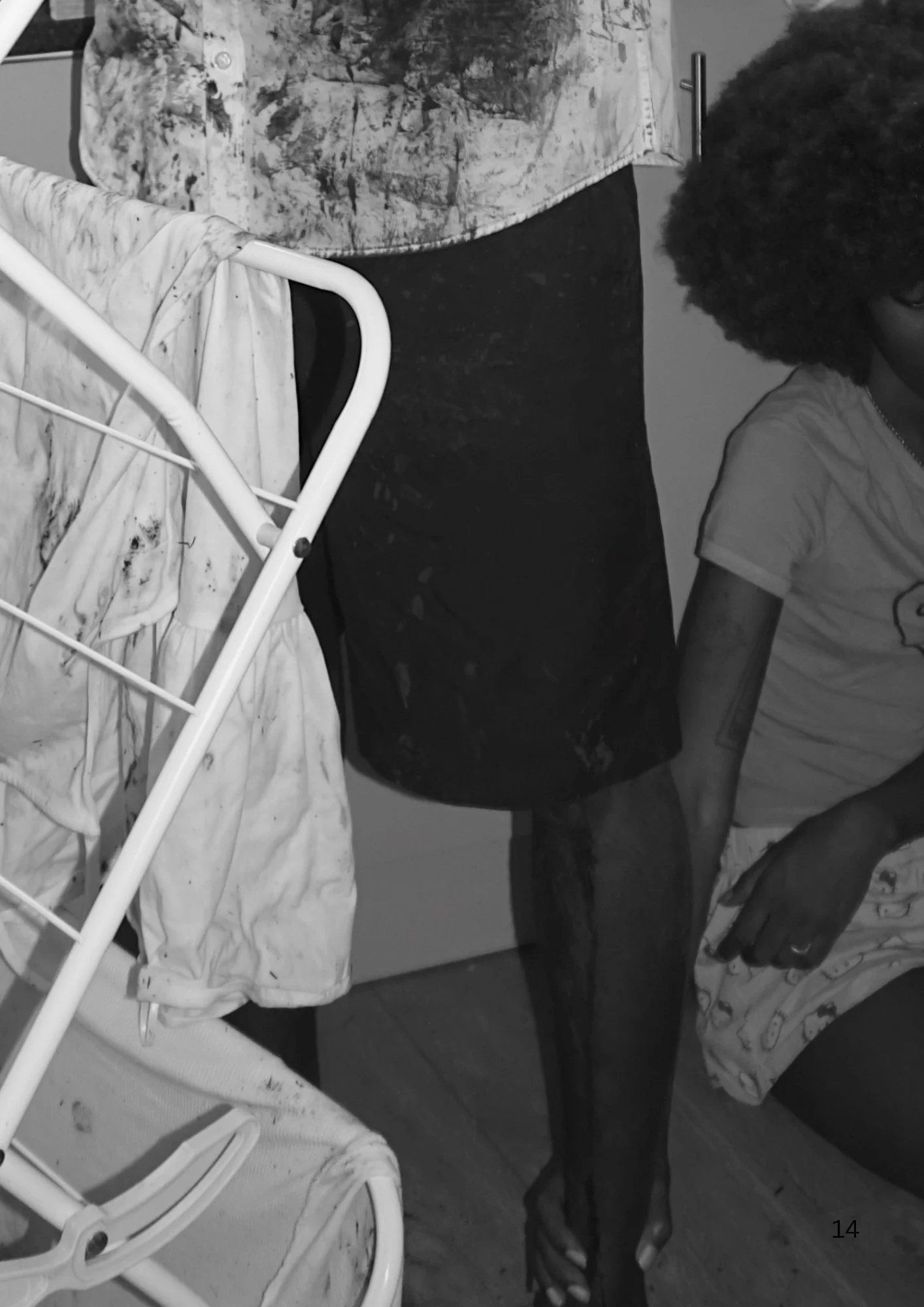 A woman with curly hair standing next to a bed and a white metal chair, wearing a light-colored T-shirt and shorts.
