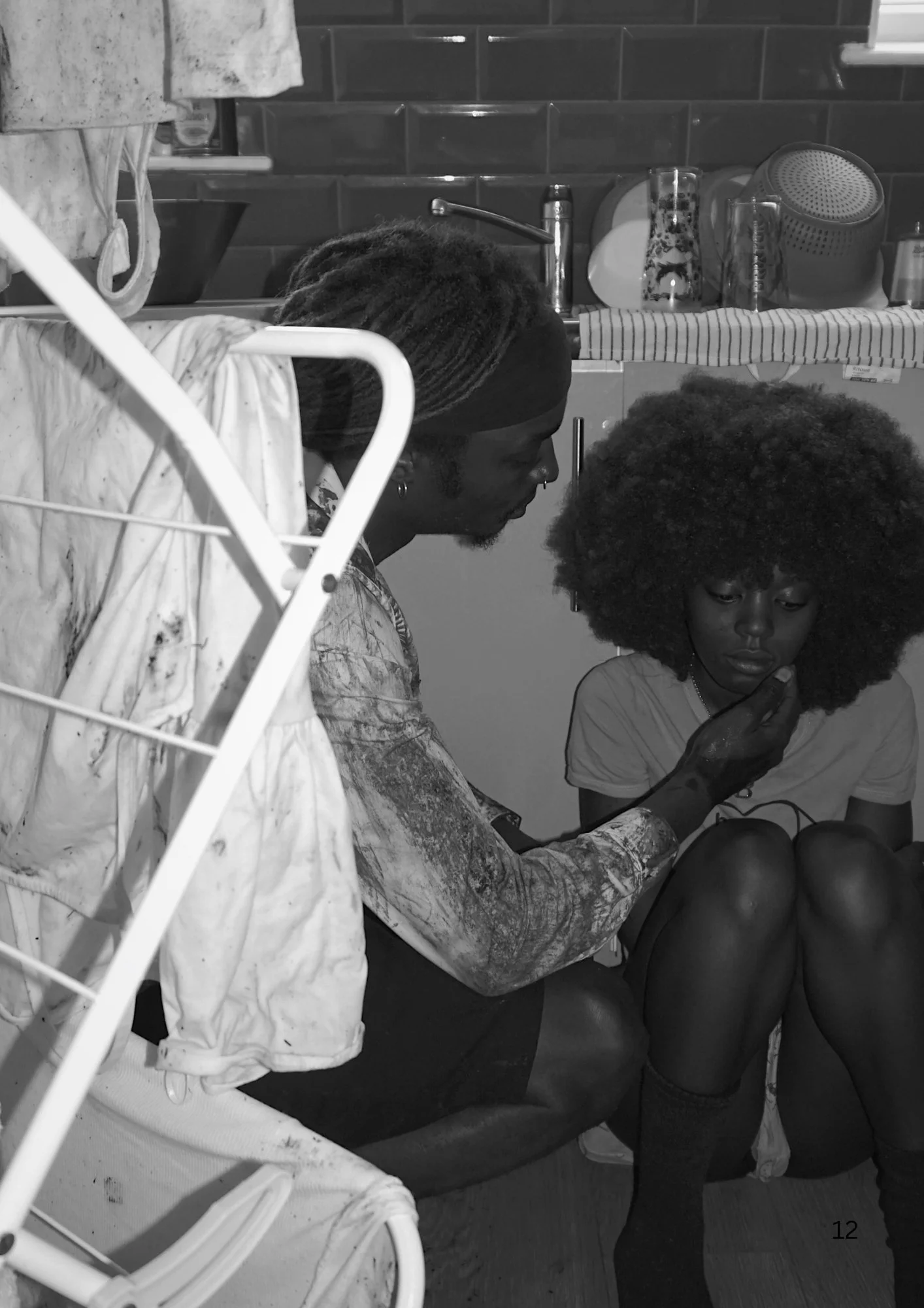 Two women, one with dreadlocks and one with voluminous curly hair, are sitting on the floor in a kitchen. The woman with curls appears to be looking at something in her lap, possibly a phone. The woman with dreadlocks is leaning forward, facing her. 