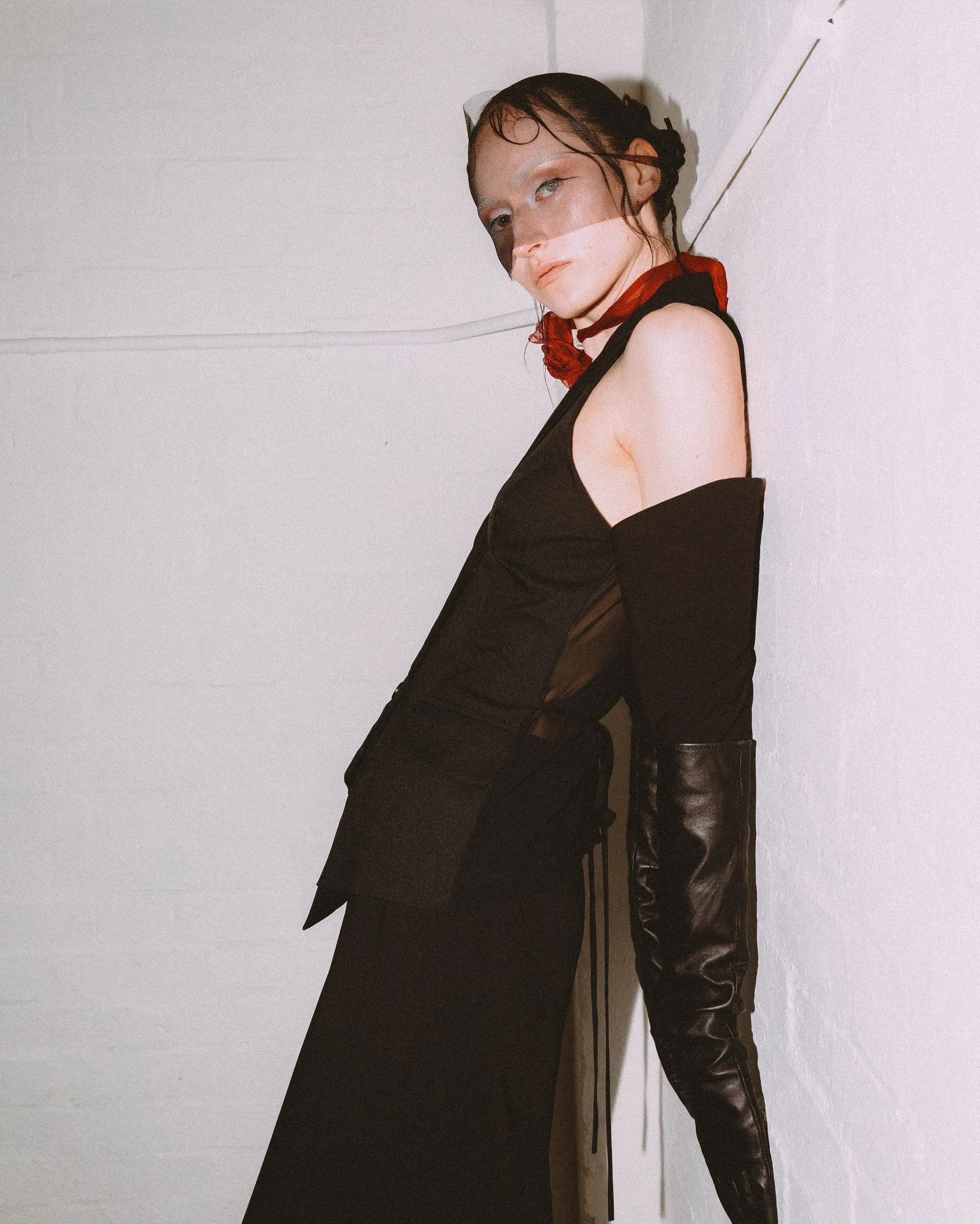 A young woman with wet hair and light makeup poses against a white wall wearing a black off-the-shoulder top and black leather pants.