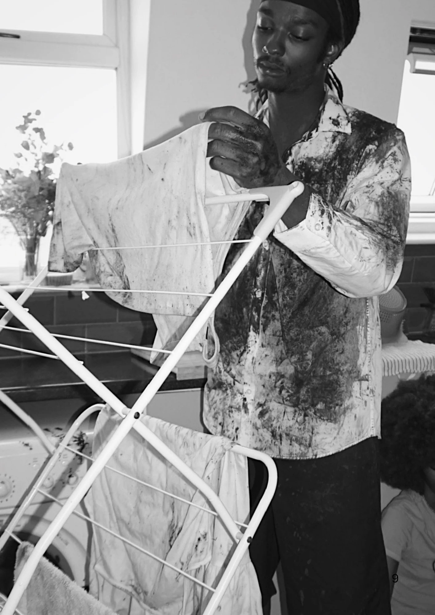 A person with dreadlocks and a nose ring, wearing a splattered shirt, is hanging laundry on a drying rack in a bright room with a window.