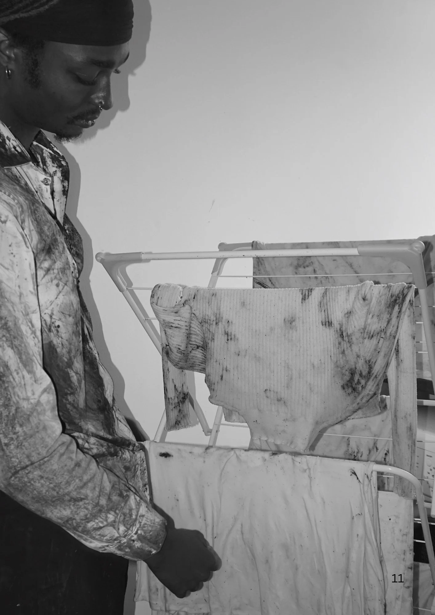 A person with dark skin, wearing a beanie and a patterned shirt, sits next to a laundry drying rack with clothes hanging on it.