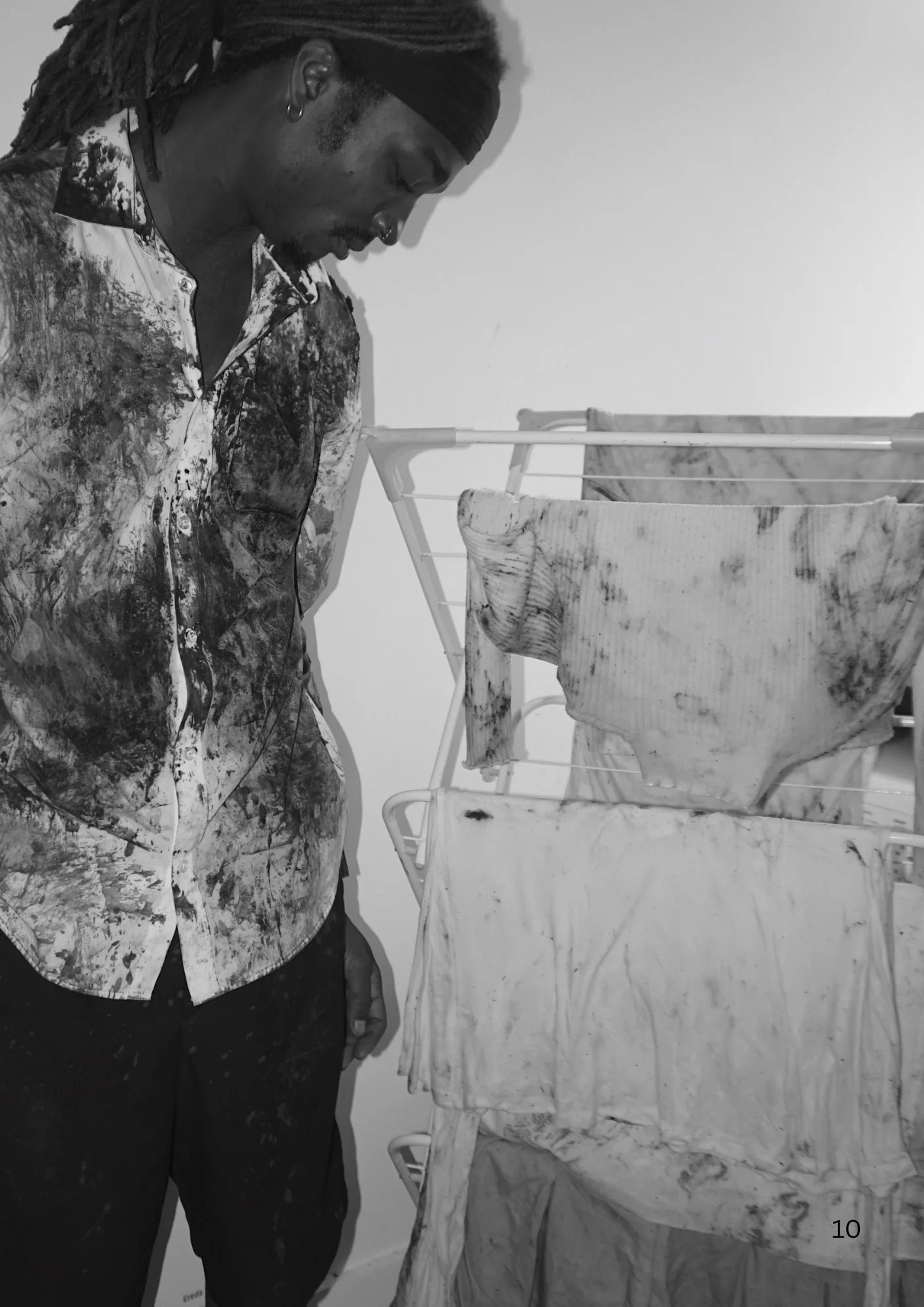 Black and white image of a man with dreadlocks wearing a patterned shirt, standing next to a clothes drying rack with various fabrics hanging on it.