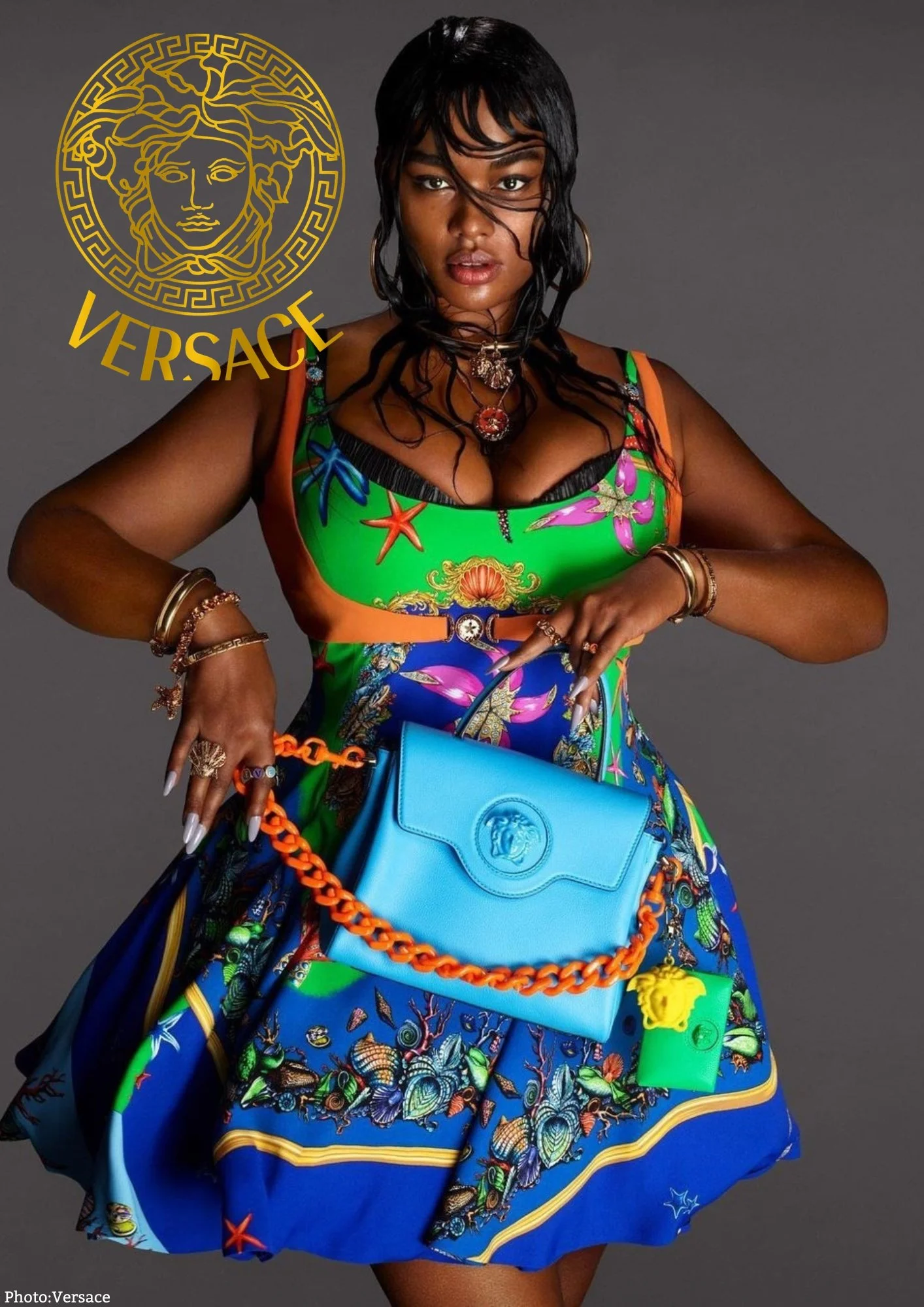 A woman with dark wavy hair and dark skin wearing a colorful printed dress, accessorized with jewelry, and holding a bright blue Versace purse with an orange chain. The Versace logo appears in the top left corner.