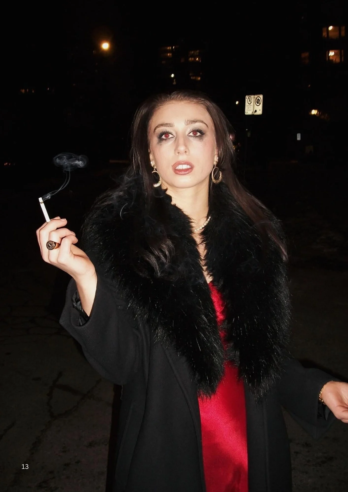 A woman with dark brown hair, wearing a black fur coat, a red dress, and gold jewelry, is standing outside at night holding a lit cigarette. She has makeup on with red eyes and lips, and is looking directly at the camera.