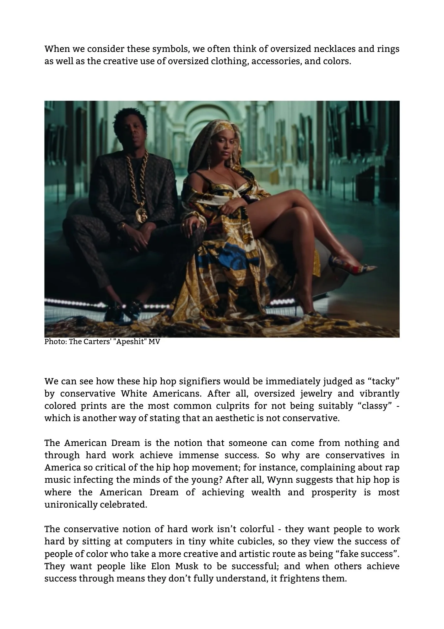 Two hip hop artists, a man and a woman, sitting on a modern sofa in a spacious, marble-floored museum or gallery, dressed in luxurious, patterned clothing with jewelry, posing confidently.