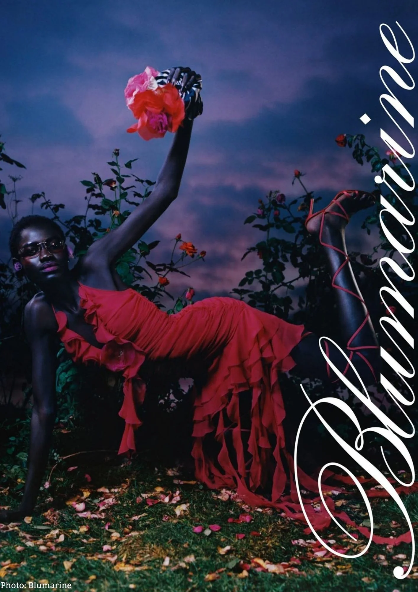 A woman wearing a red dress and glasses is lying on the ground amidst fallen leaves, holding a bouquet of pink and red flowers in the air. The sky behind her is a deep blue with purple hues, and the scene has a dreamy, artistic vibe.
