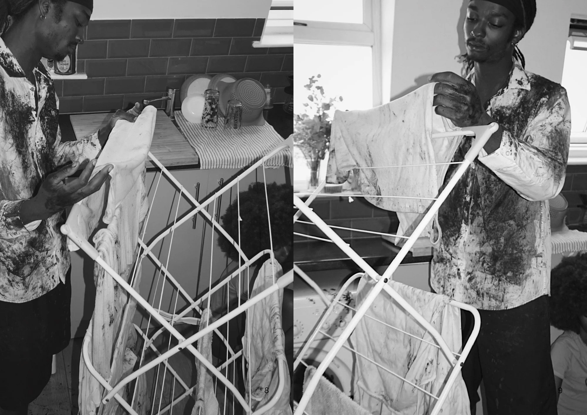 A black and white image of a man with dreadlocks wearing a patterned shirt, drying a towel on a white drying rack in a kitchen. The kitchen has a brick backsplash, a window, and various dishes on a dish drying mat.