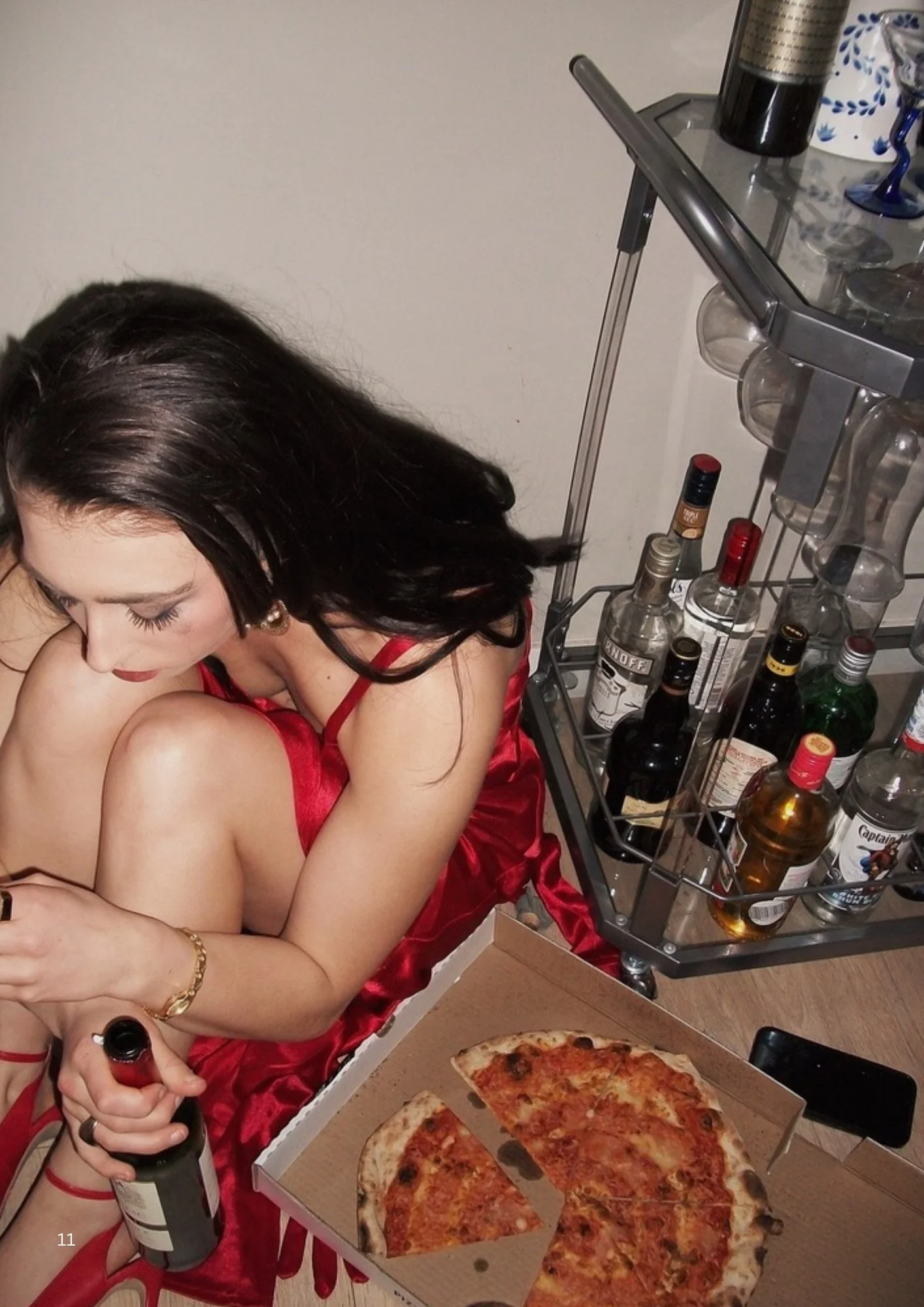 A woman with dark hair in a red dress sitting on the floor next to a table with pizza, holding a bottle of wine, with liquor bottles on a serving cart nearby.