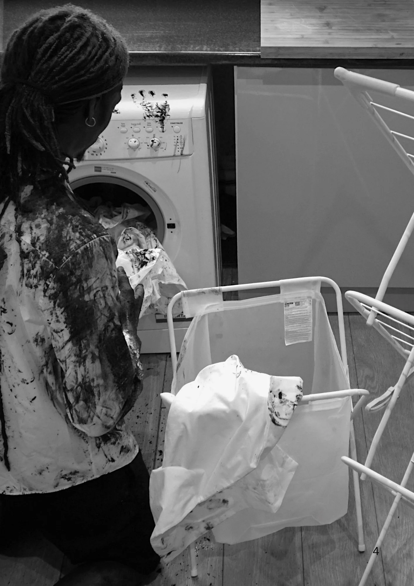 A person with braided hair wearing a dark patterned shirt is standing in front of a washing machine, which has paint splatters on it, in a laundry room. There are laundry hampers and chairs nearby.