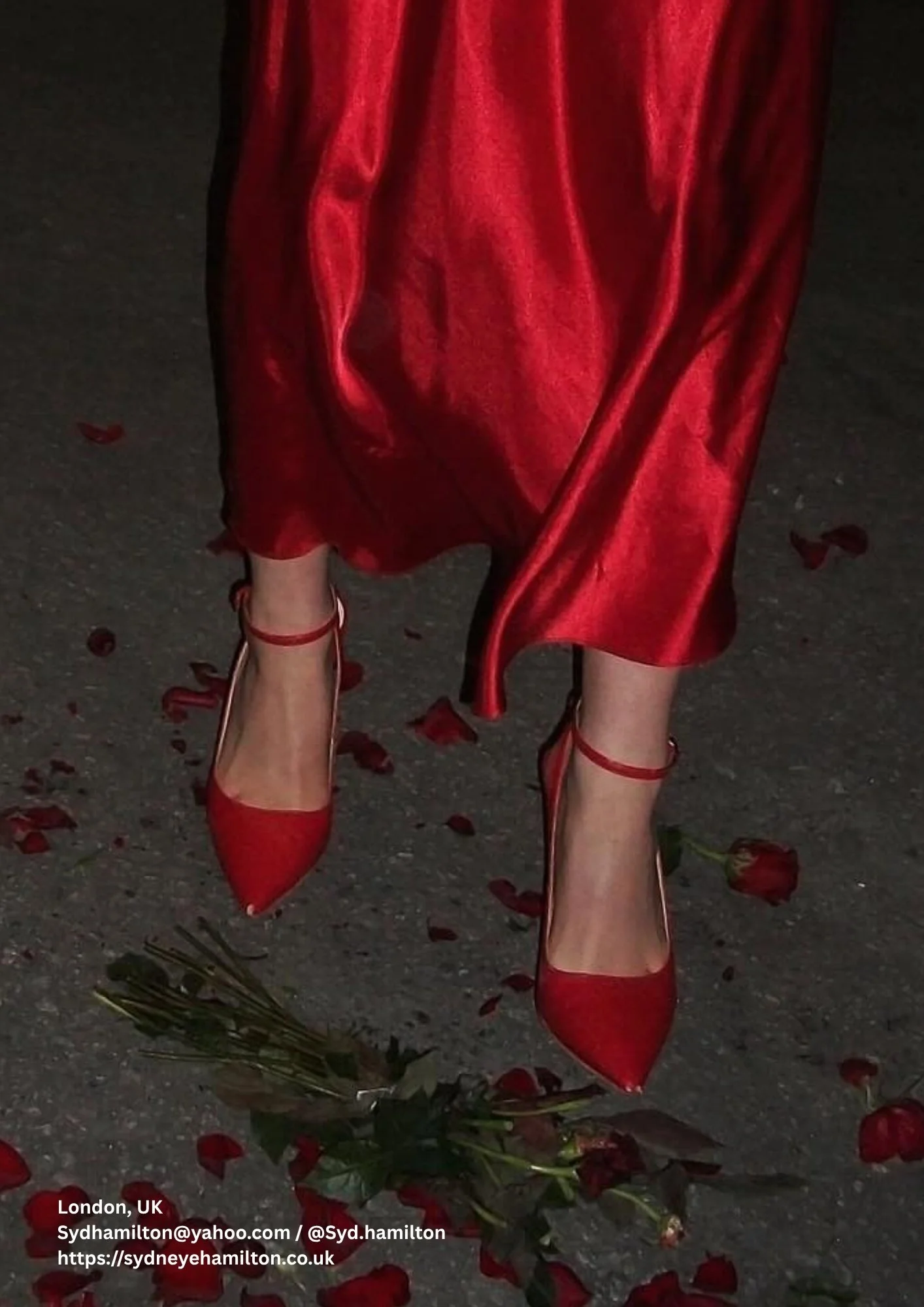 A person wearing red high-heeled shoes and a matching red satin skirt walking on a street with rose petals and leaves scattered on the ground.