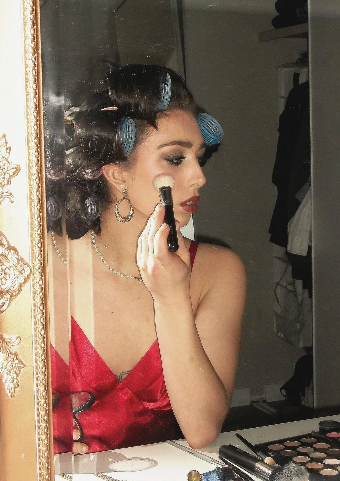 A woman with dark hair in rollers is applying makeup using a brush in front of a mirror.