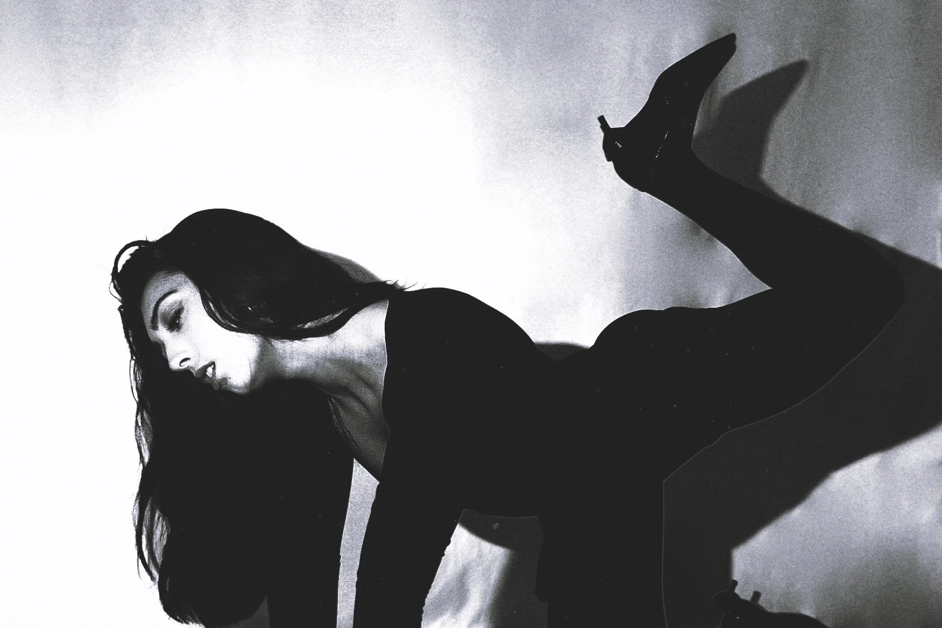 A black and white photo of a woman with long dark hair, dressed in black, sitting with her face turned to the side and one leg raised, casting a shadow on the wall behind her.