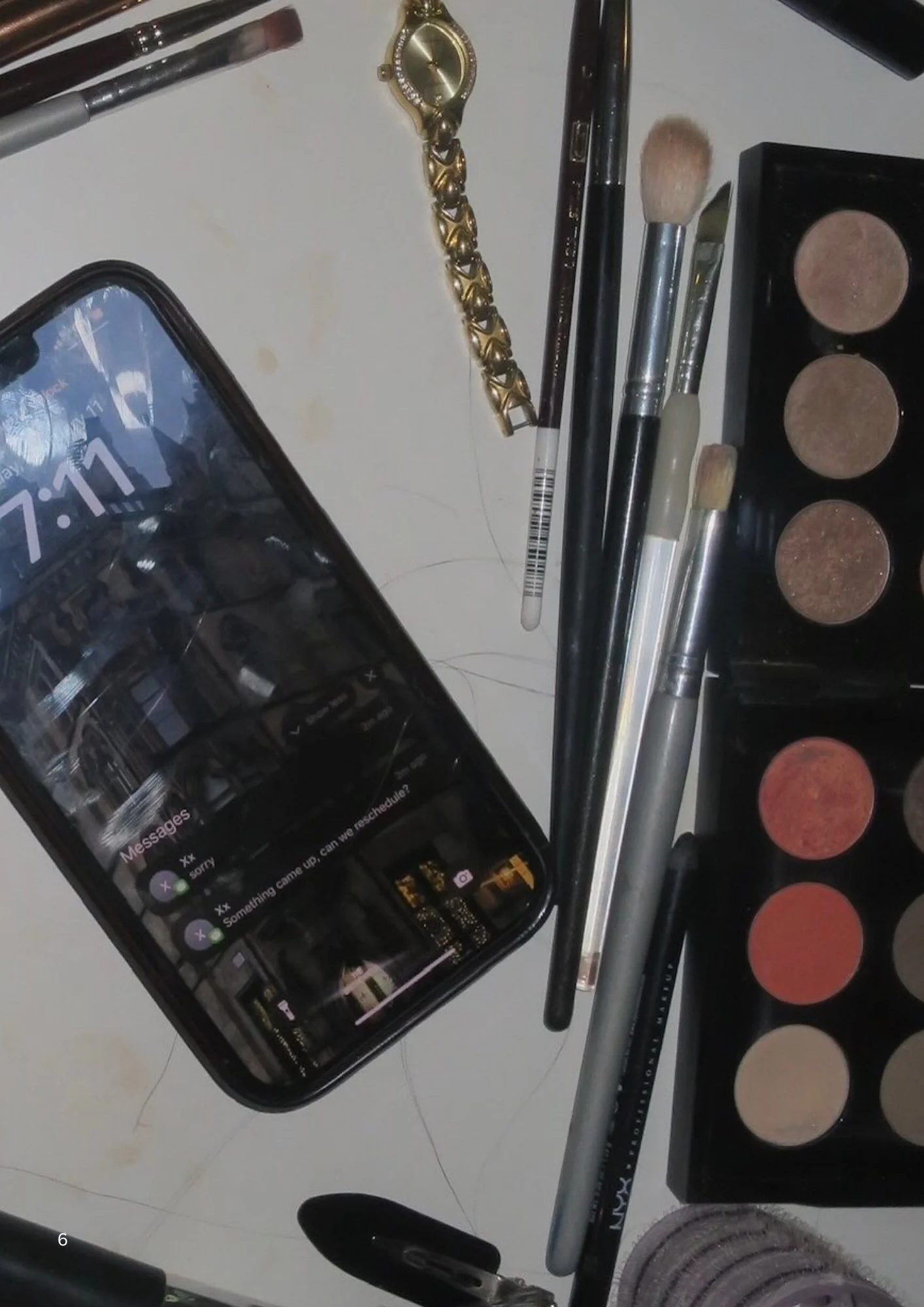 Makeup brushes, eyeshadow palette, watch, phone, and a ring on a desk.