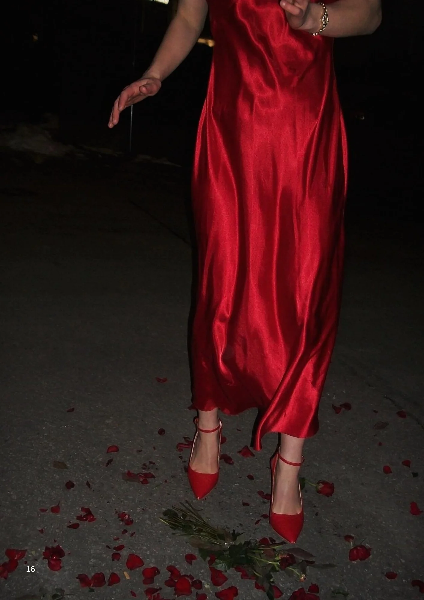A woman in a long, red satin dress and red high heels walking on a street scattered with rose petals, with a bouquet at her feet.