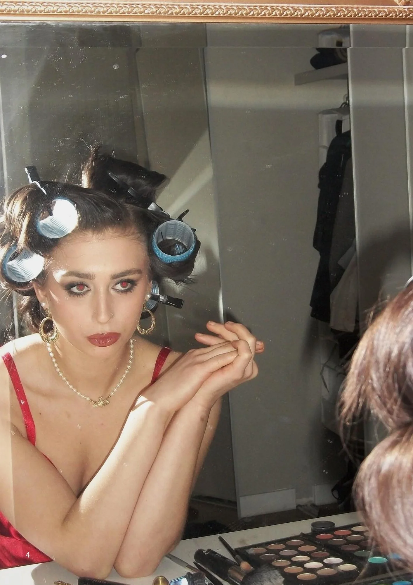 A woman with curled hair in rollers, makeup, and jewelry sitting in front of a mirror, applying makeup with an eyeshadow palette and brushes visible on the table.