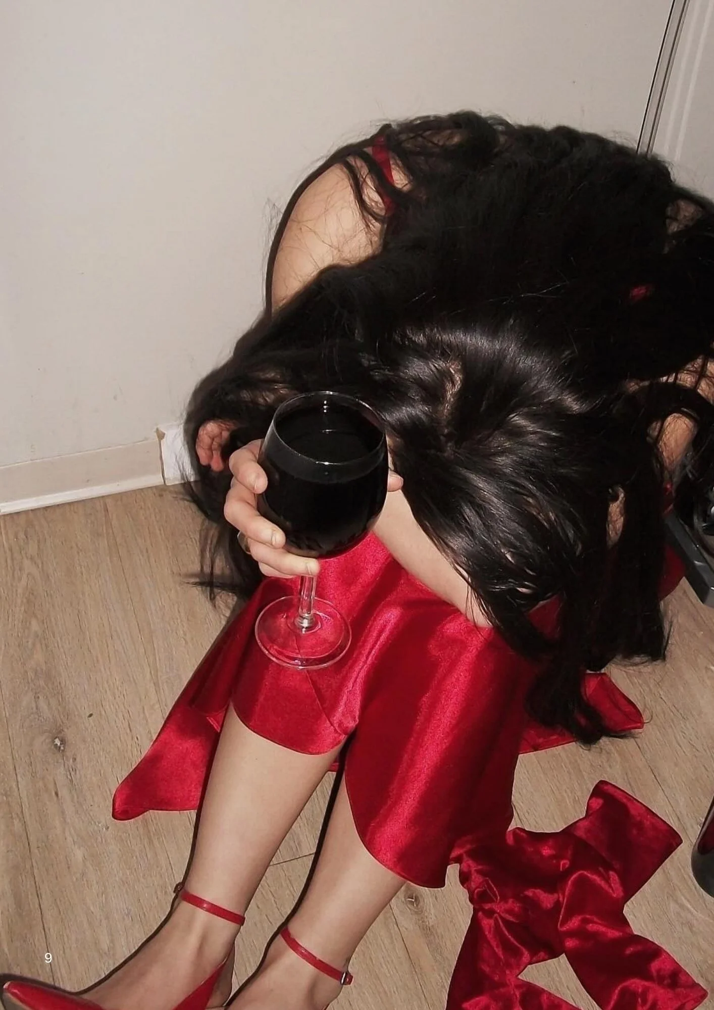 A woman with long black hair wearing red shoes, red dress, and red ankle strap heels, sitting on the floor and holding a glass of red wine.
