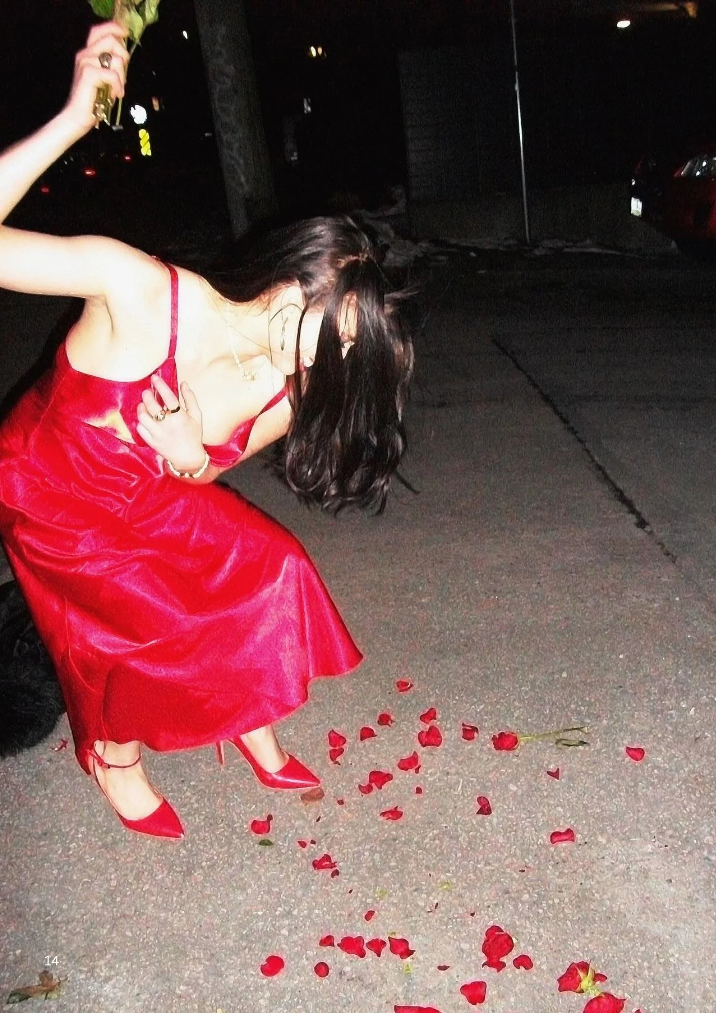 A woman in a red dress and high-heeled shoes is kneeling or bending over on a street at night, with rose petals scattered on the ground around her.