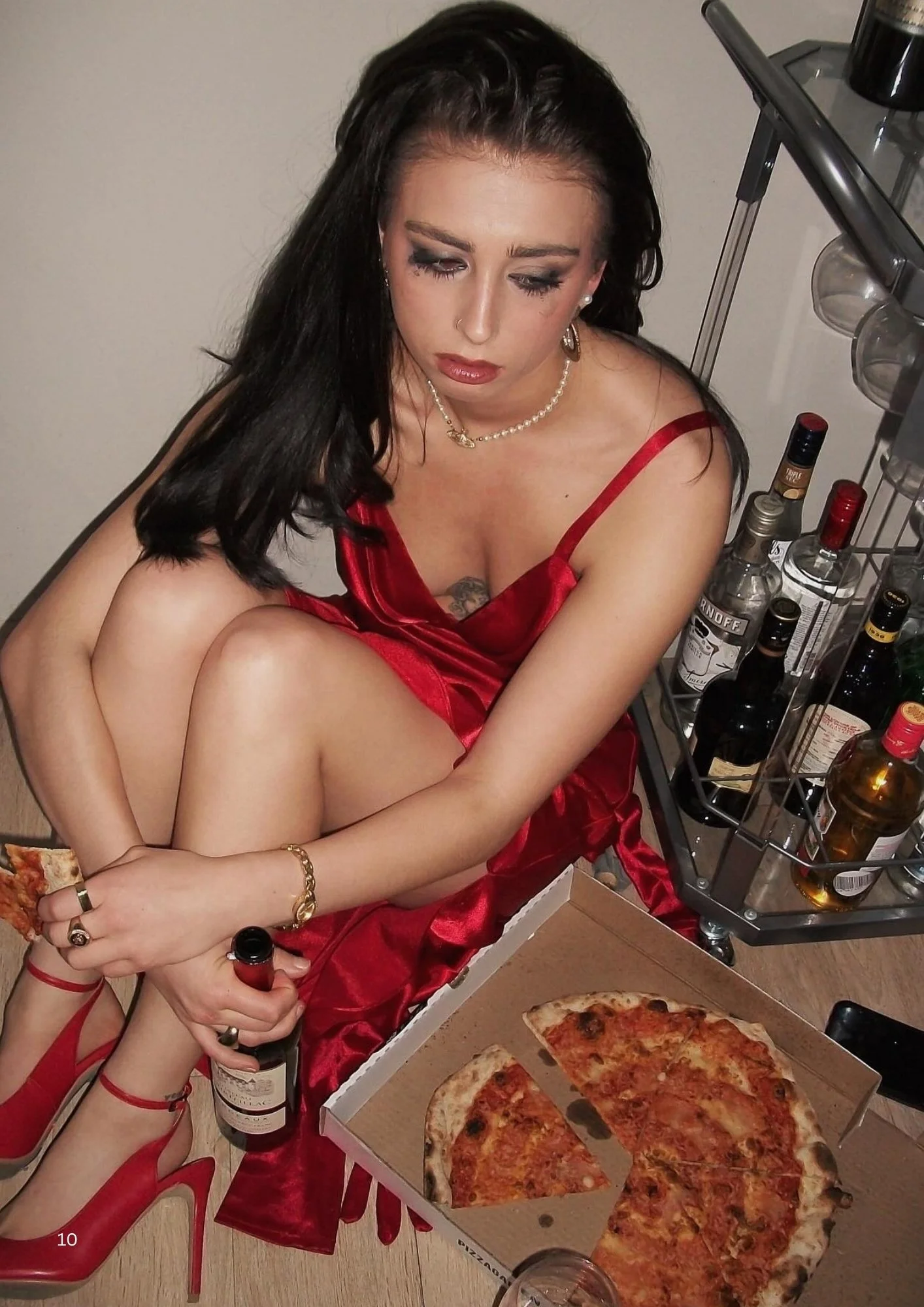 A woman in a red dress sitting on a red chair, with multiple bottles of alcohol and a pizza box with several slices of pizza on a table nearby.