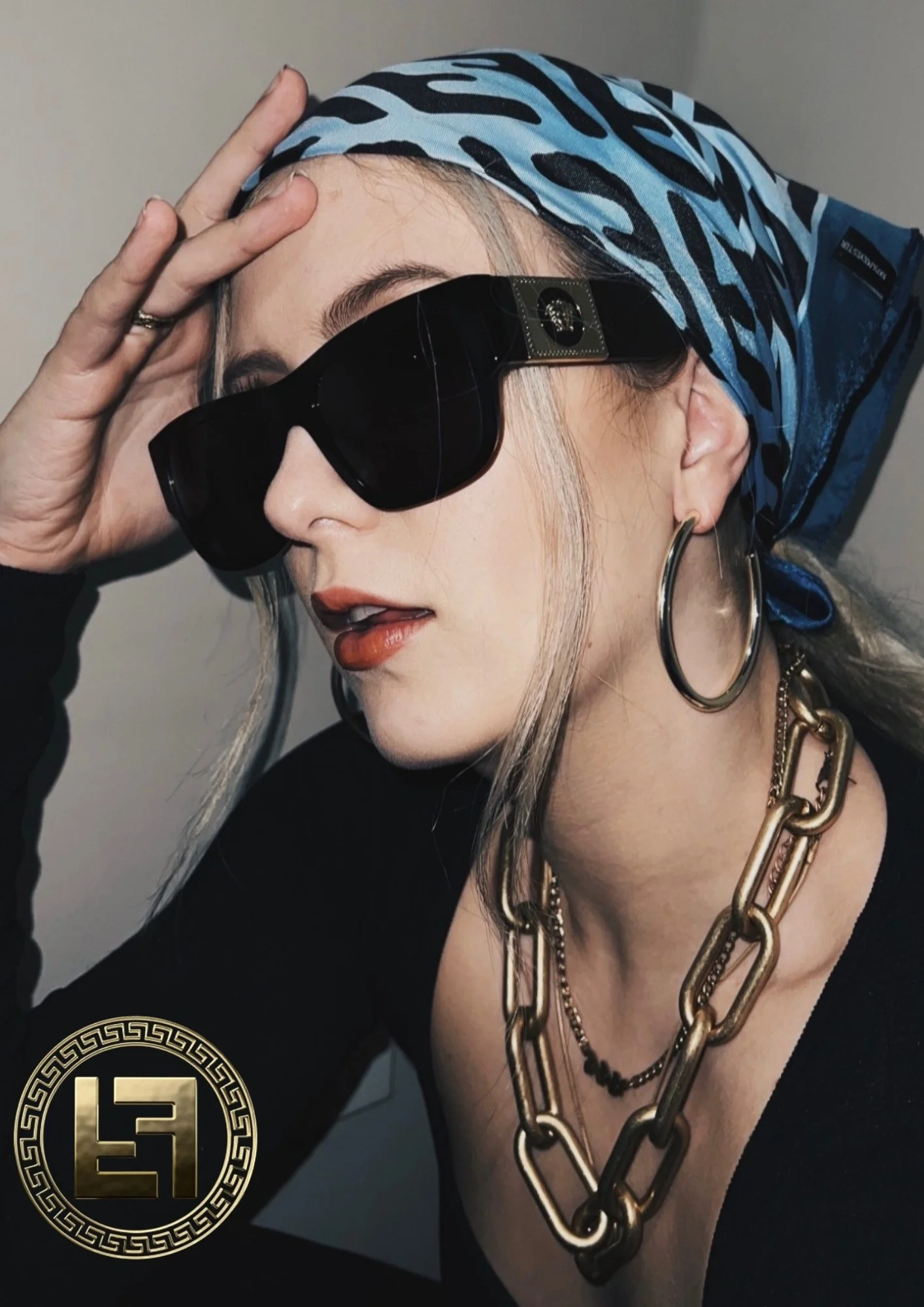 A woman wearing large hoop earrings, a blue and black patterned headscarf, black sunglasses, and layered gold chain necklaces, holding her forehead with one hand.