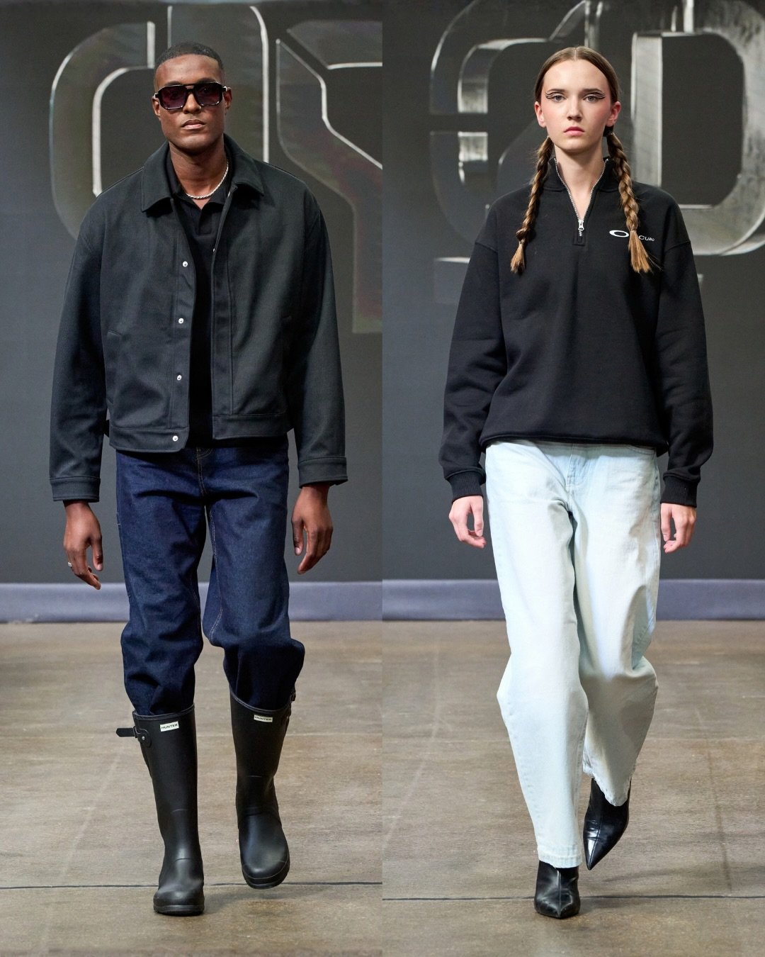 Two fashion models walking on a runway, wearing casual streetwear. The male model is dressed in a black jacket, black sunglasses, dark jeans, and black boots. The female model is wearing a black pullover with a zipper, light-colored jeans, and black pointed boots. They are walking in front of a dark backdrop with metallic block-style lettering.
