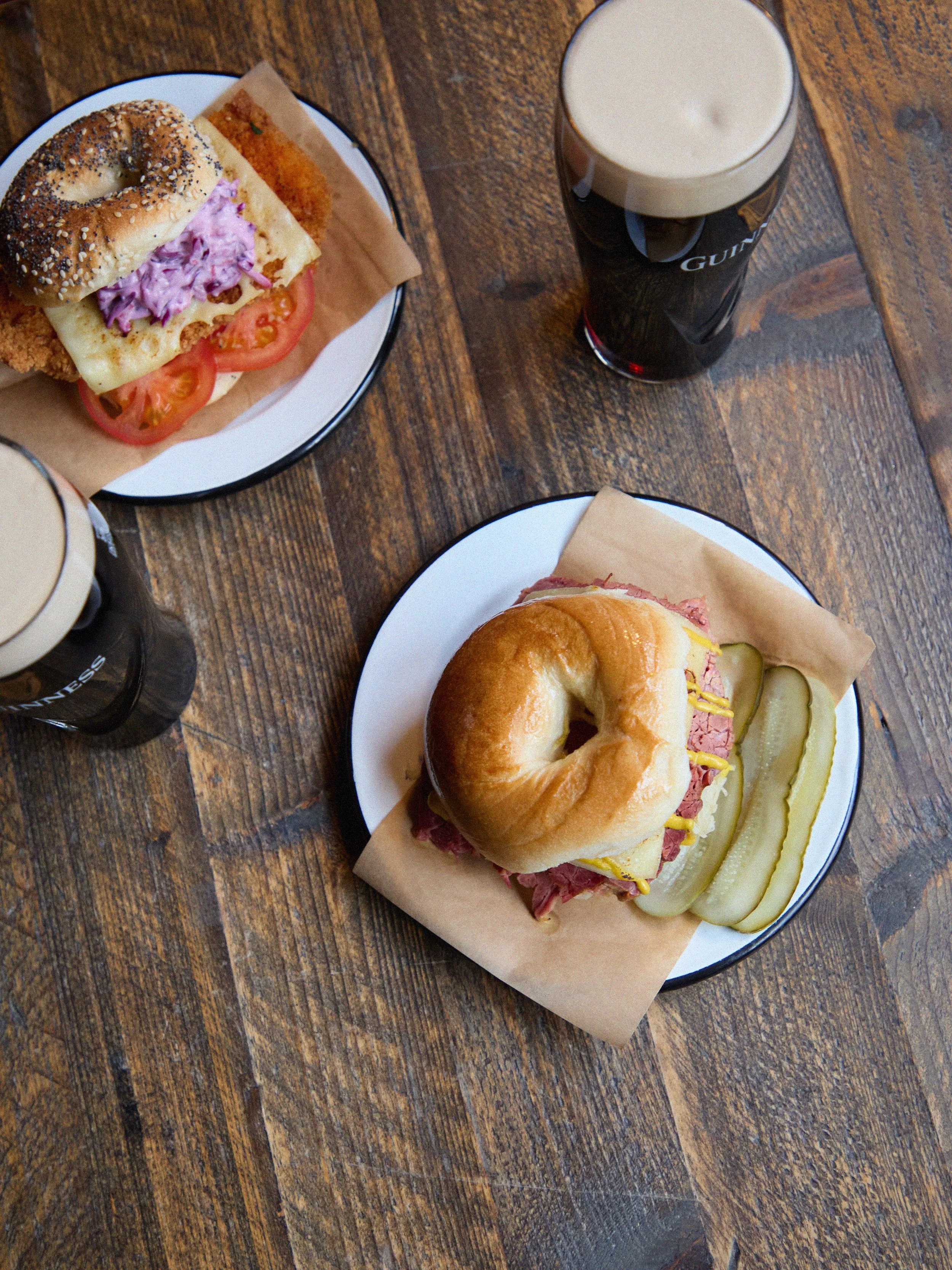 Two bagel sandwiches on plates with pints of Guinness on a wooden table.