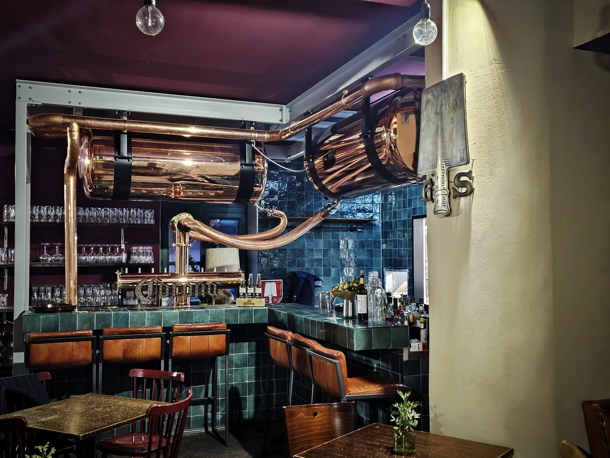 Innenansicht einer Bar mit kupferner Zapfanlage, grünen Fliesen, Holzstühlen und einer Bar mit Gläsern und Flaschen.