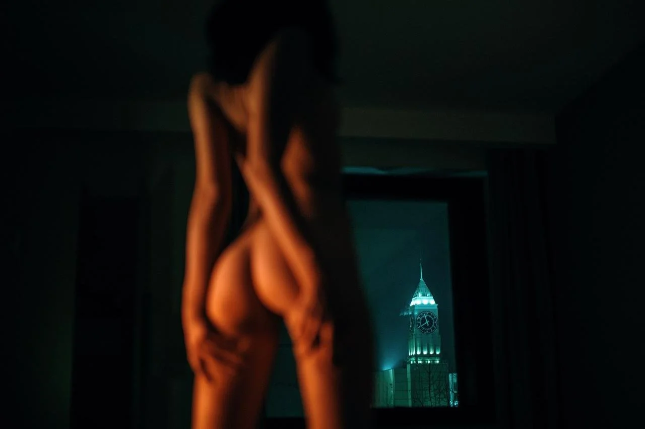 Une silhouette d'une personne nue qui se tient dans une pièce sombre, avec une vue sur une tour illuminée au fond, probablement une horloge, à travers une fenêtre.